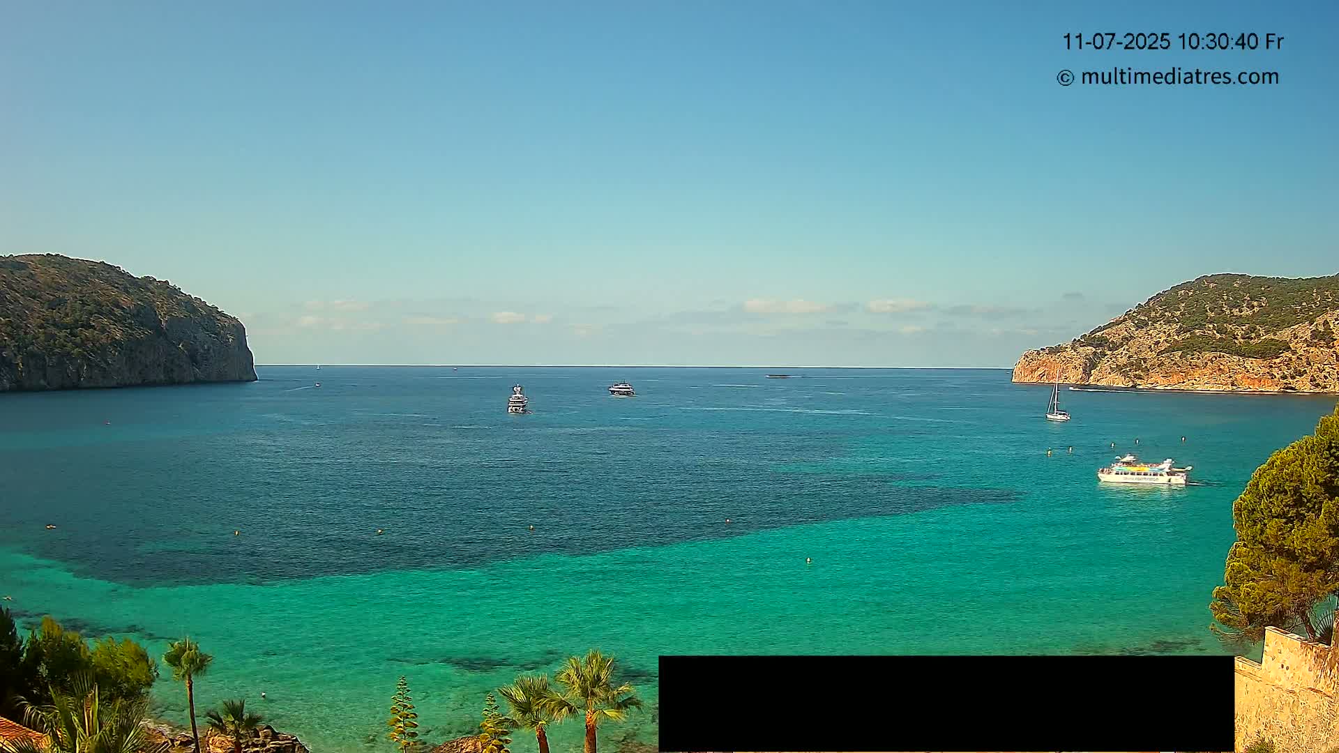 A calm, sunny day reveals a picturesque bay with clear, turquoise water, surrounded by rocky hills and several boats.