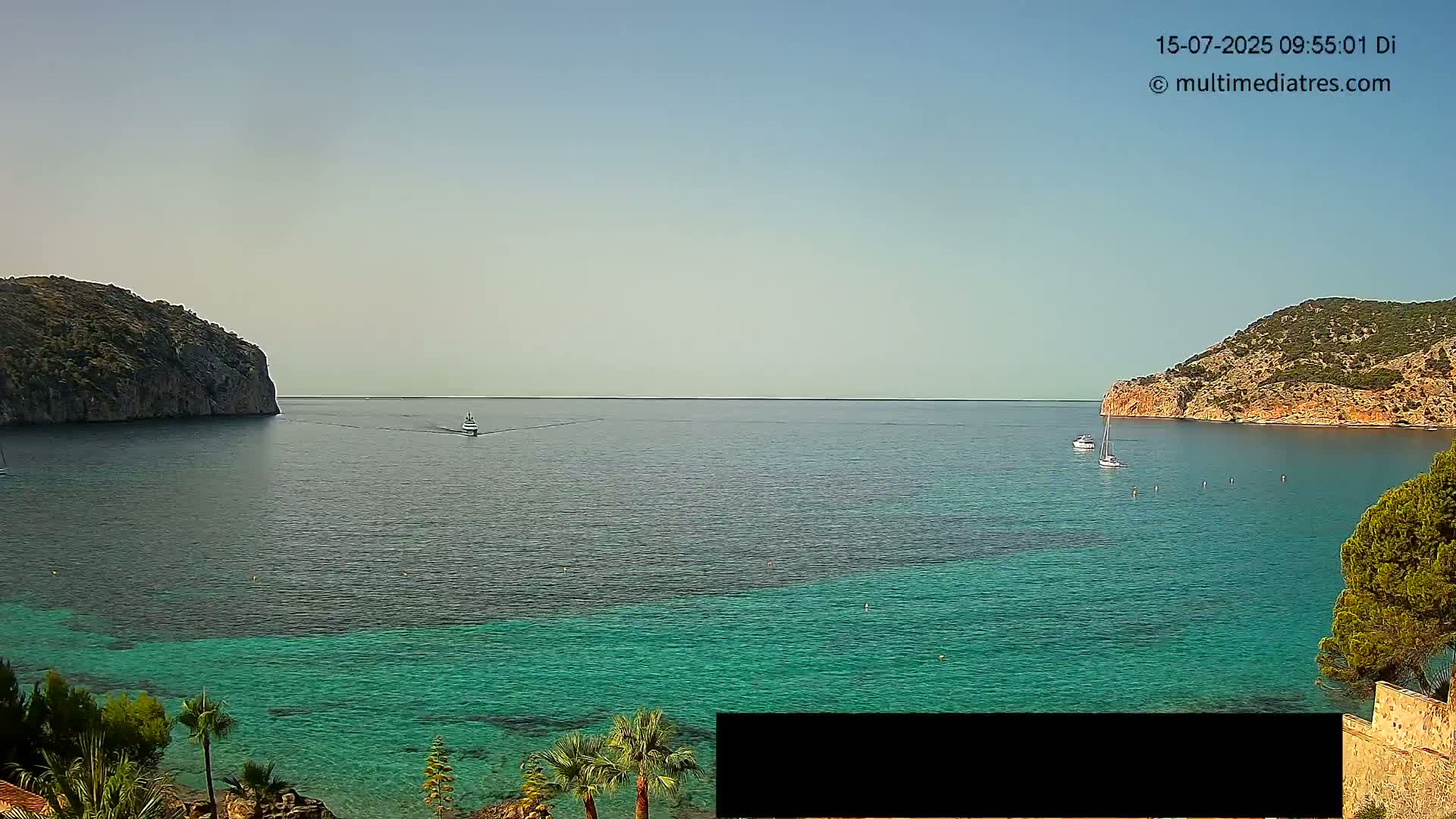 A clear, sunny day reveals a tranquil cove with calm, teal water, two small boats, and rocky hillsides on either side.