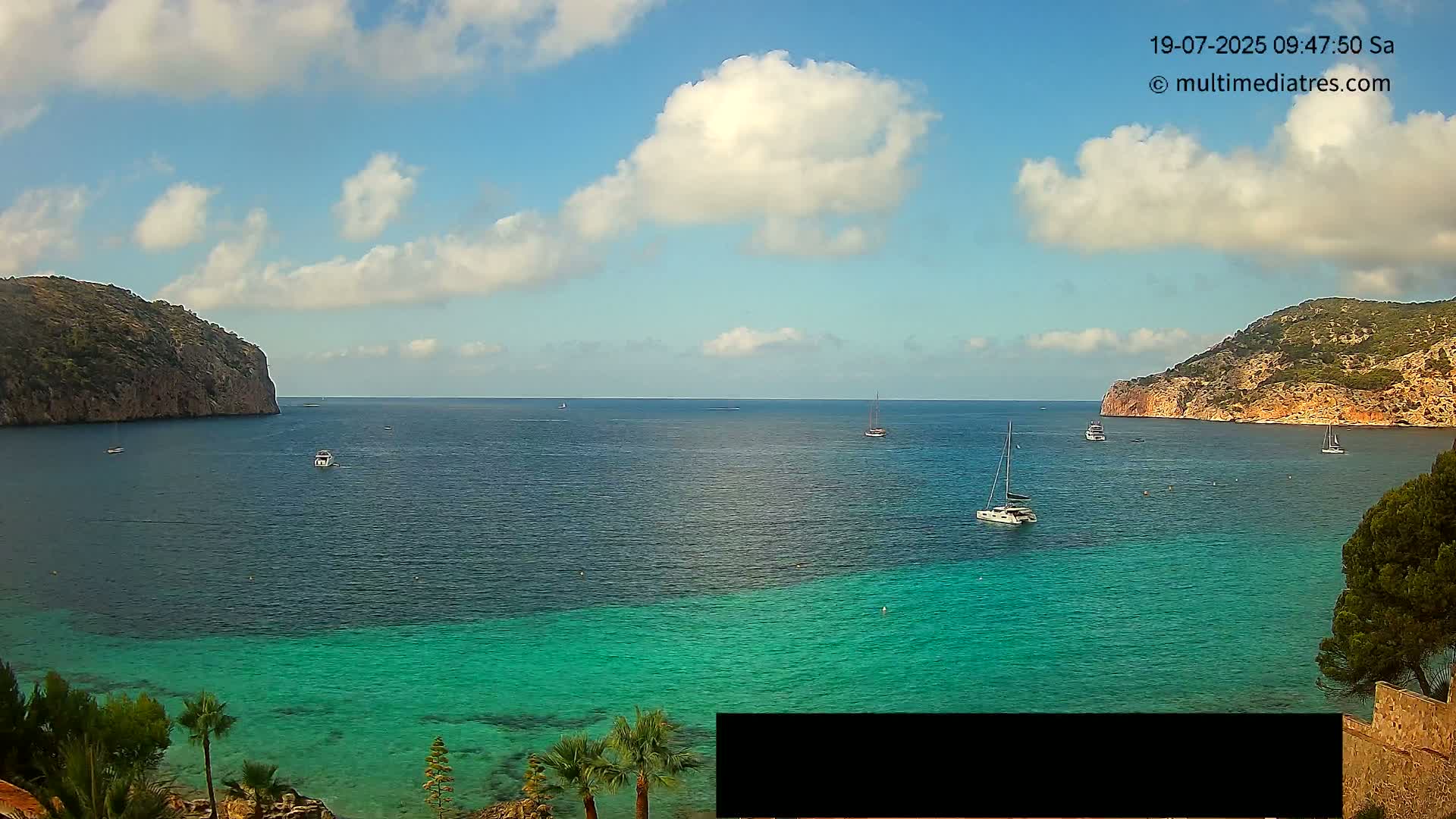 A partly cloudy day reveals a tranquil bay with turquoise water, several boats, and steep, rocky hillsides on either side.