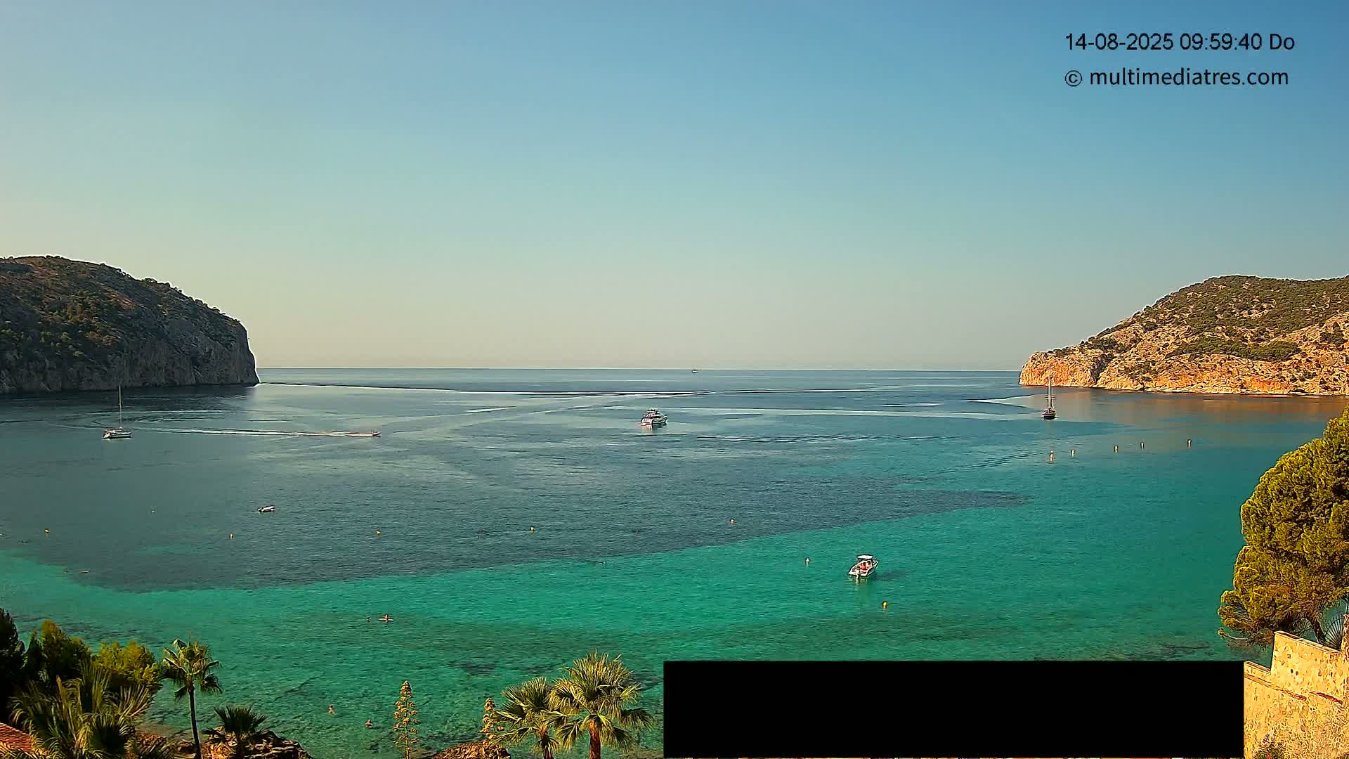 A calm, clear day reveals a tranquil cove with turquoise water, several boats, and rocky hills on either side.