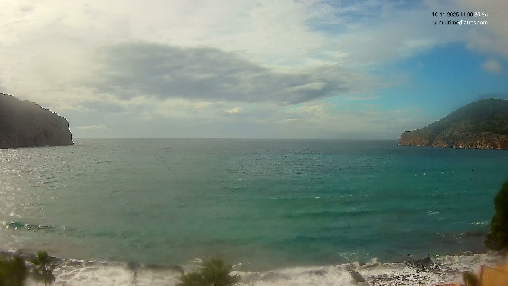 A scenic coastal bay features choppy turquoise-green water with foamy waves breaking on a rocky shore, framed by steep, green-covered cliffs on either side, all under a partly cloudy sky with patches of blue.