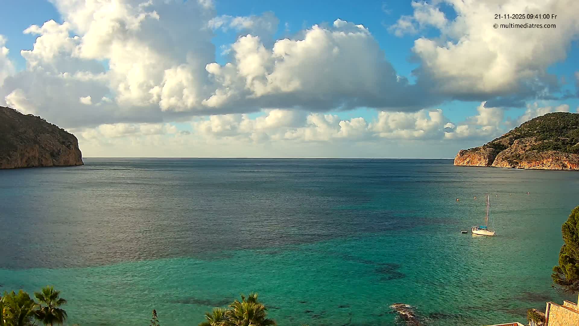 A serene coastal bay with clear turquoise water and a white sailboat anchored, framed by rocky, vegetated cliffs, is pictured under a partially cloudy blue sky with bright sunshine.