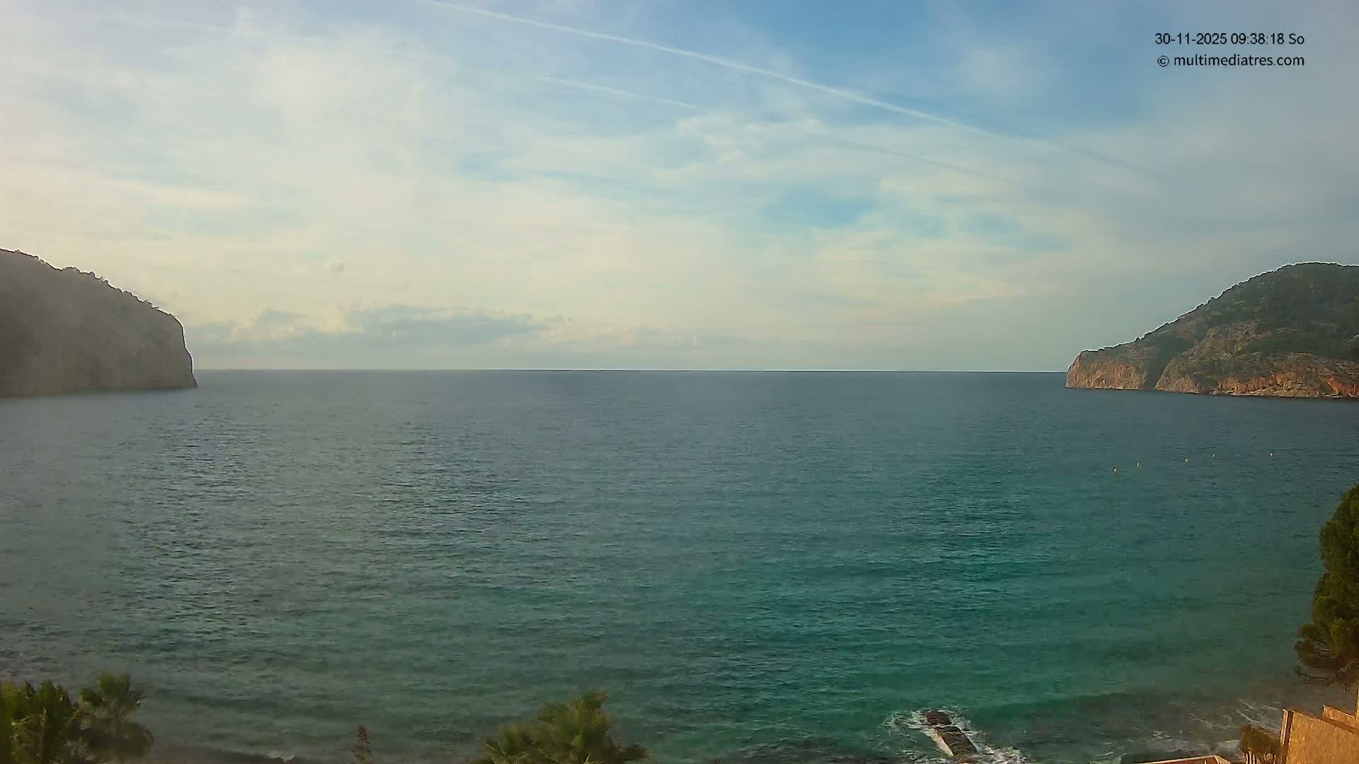 A wide view shows a calm blue-green sea nestled between rugged, vegetated cliffs on both sides, with some green plants in the foreground, all beneath a partly cloudy sky.