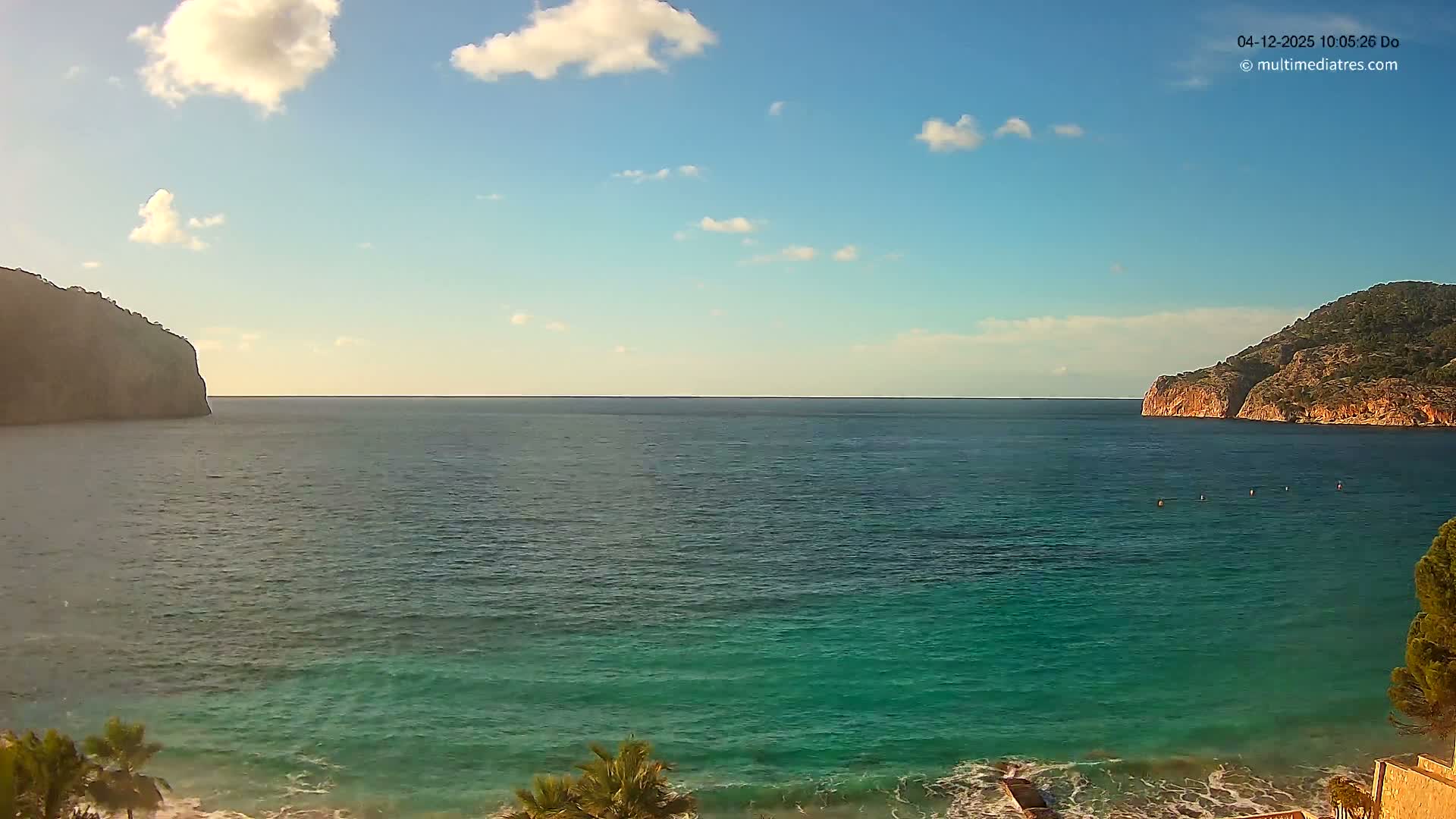 A beautiful bay with turquoise water and gentle waves is framed by rocky, vegetated cliffs on either side, all bathed in sunlight under a clear blue sky dotted with fluffy white clouds.