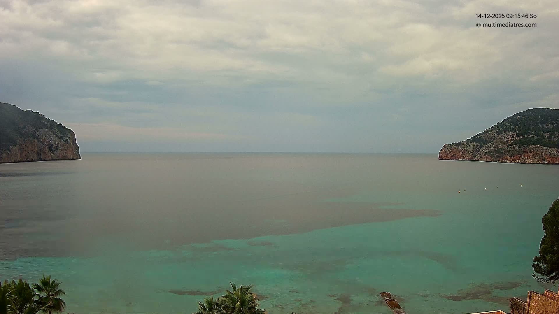 A beautiful bay with turquoise water and gentle waves is framed by rocky, vegetated cliffs on either side, all bathed in sunlight under a clear blue sky dotted with fluffy white clouds.