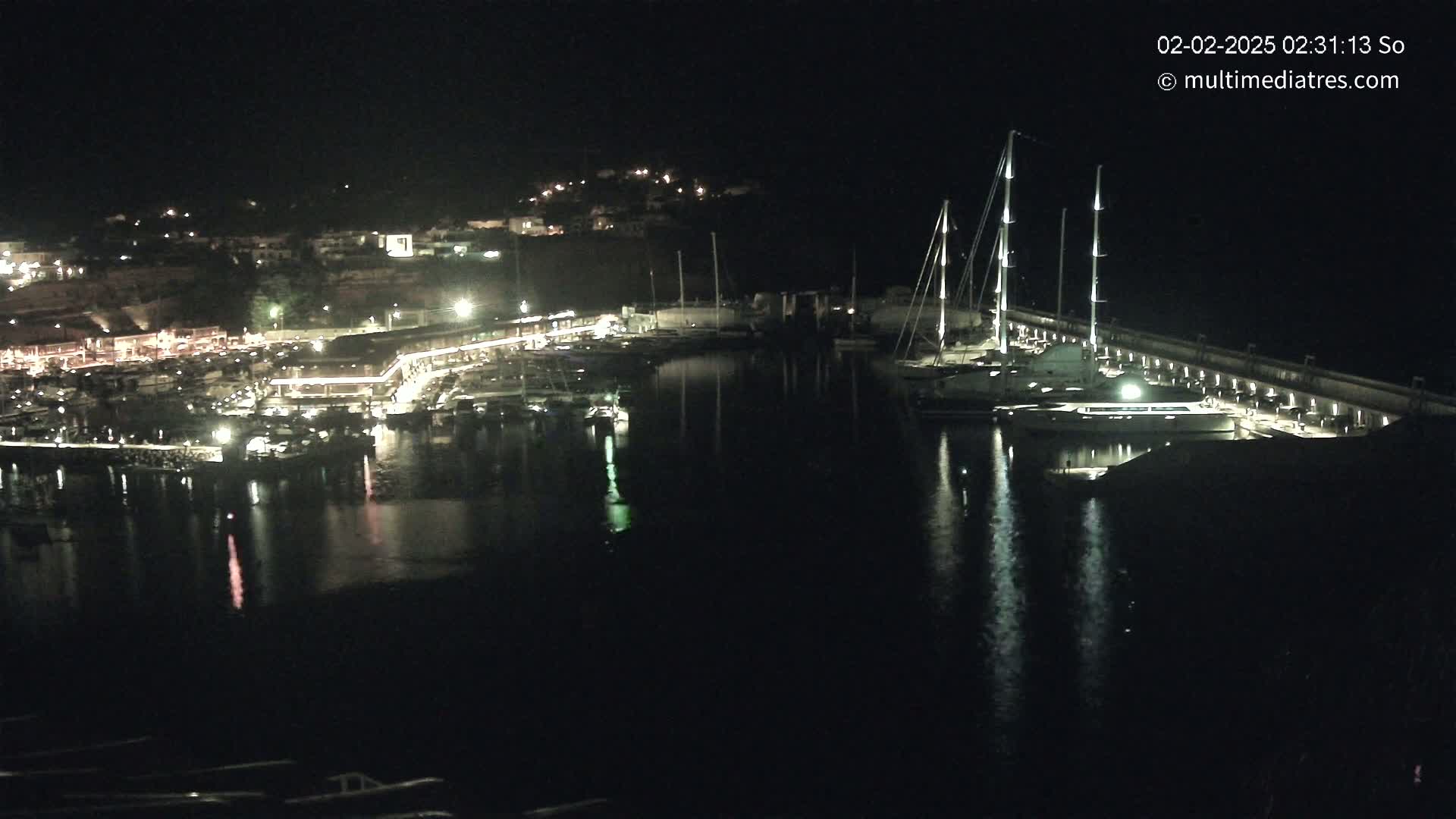 A nighttime view of a marina filled with boats, illuminated by lights against a dark, calm body of water.