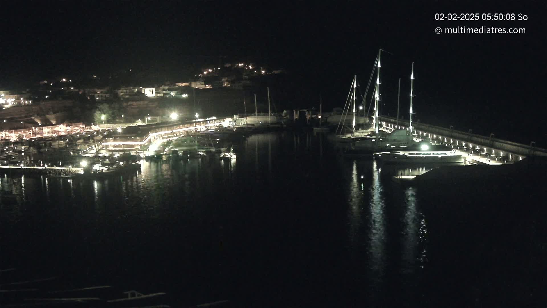 A nighttime view of a harbor filled with boats and yachts, illuminated by lights along a waterfront lined with buildings.