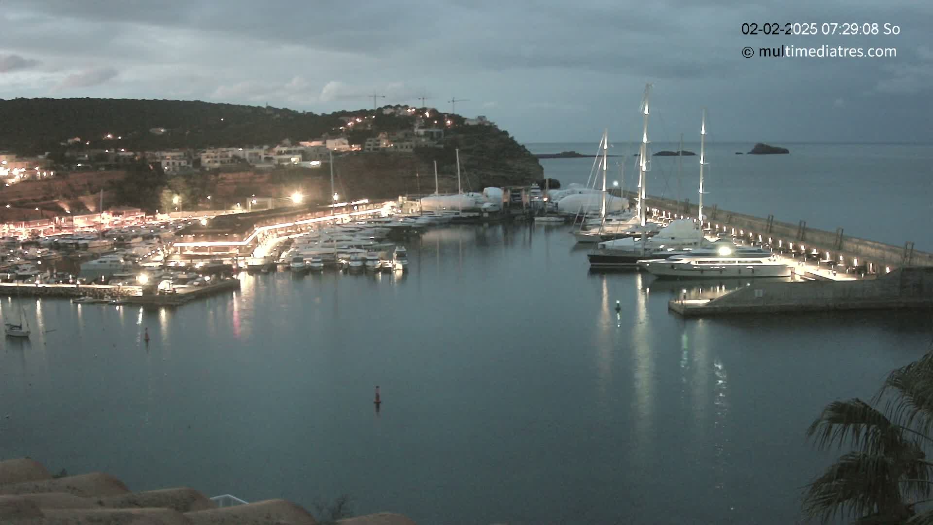 A marina filled with yachts and sailboats is illuminated at night under a cloudy sky.