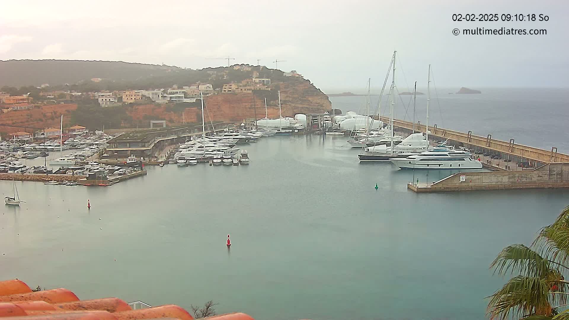 A marina filled with numerous yachts and boats is nestled against a hillside under an overcast sky.