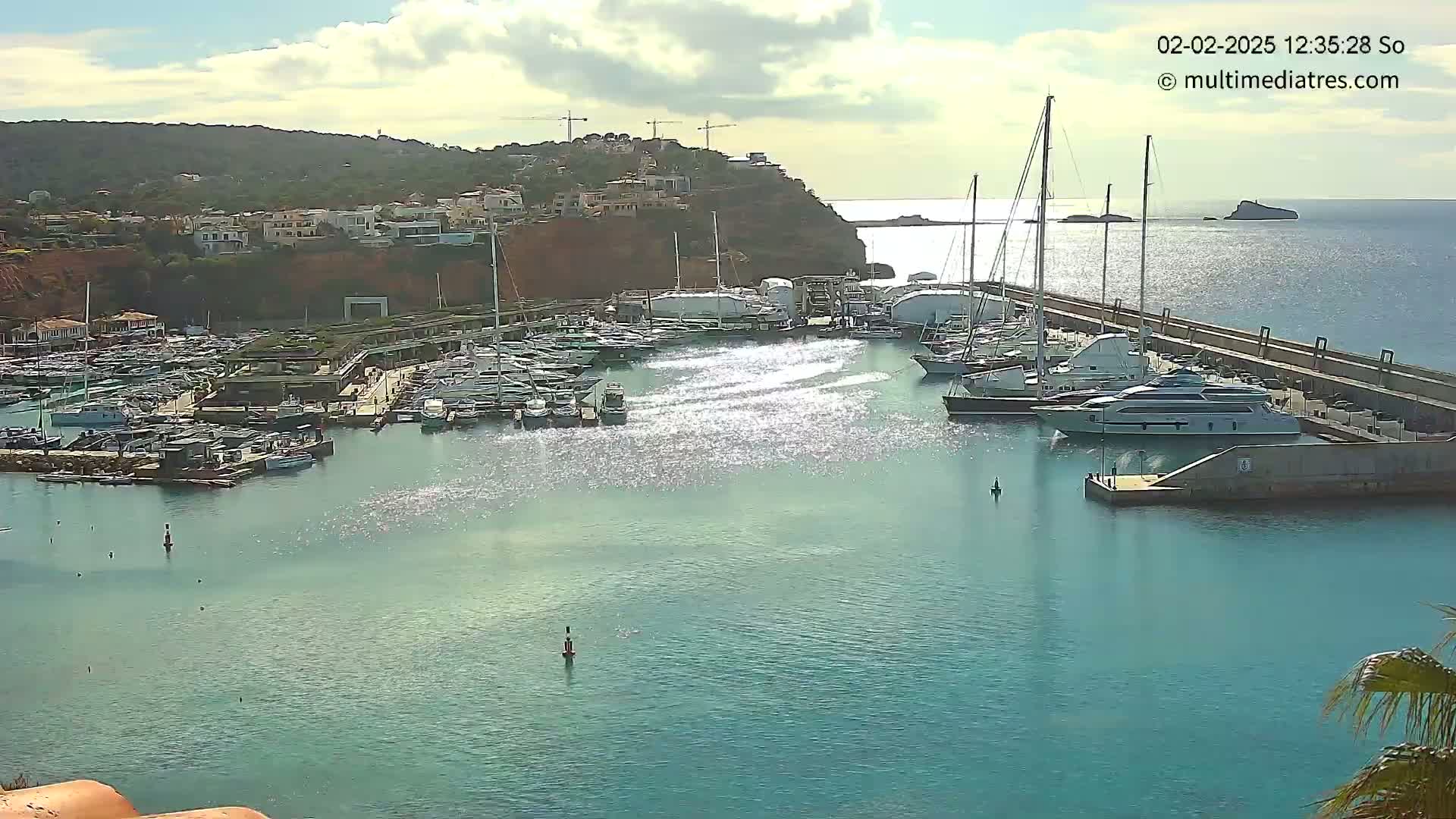 A marina filled with numerous boats and yachts is nestled beside a rocky hillside under a partly cloudy sky.