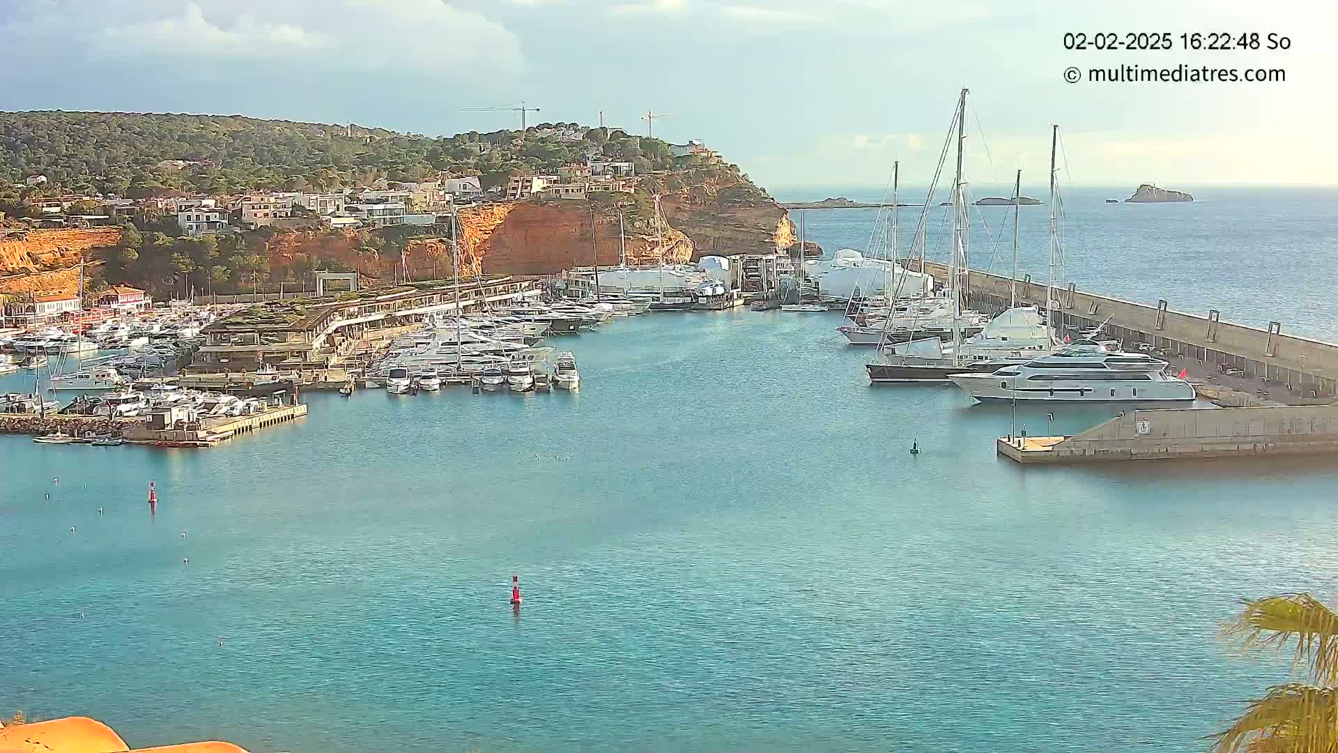 A marina filled with yachts and sailboats is situated next to a red-cliff hillside under a partly cloudy sky.