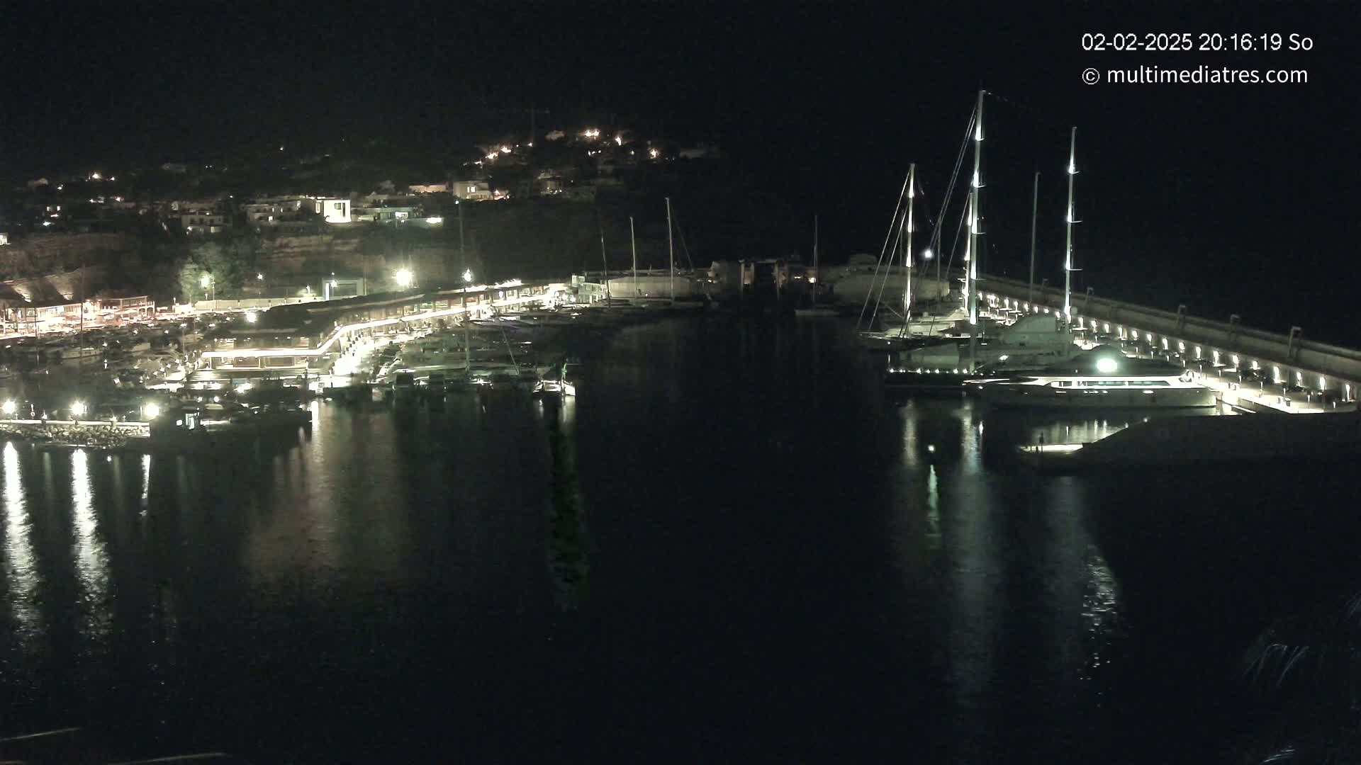 A nighttime view of a marina filled with boats, illuminated buildings along the waterfront, and calm, dark water reflecting the lights.