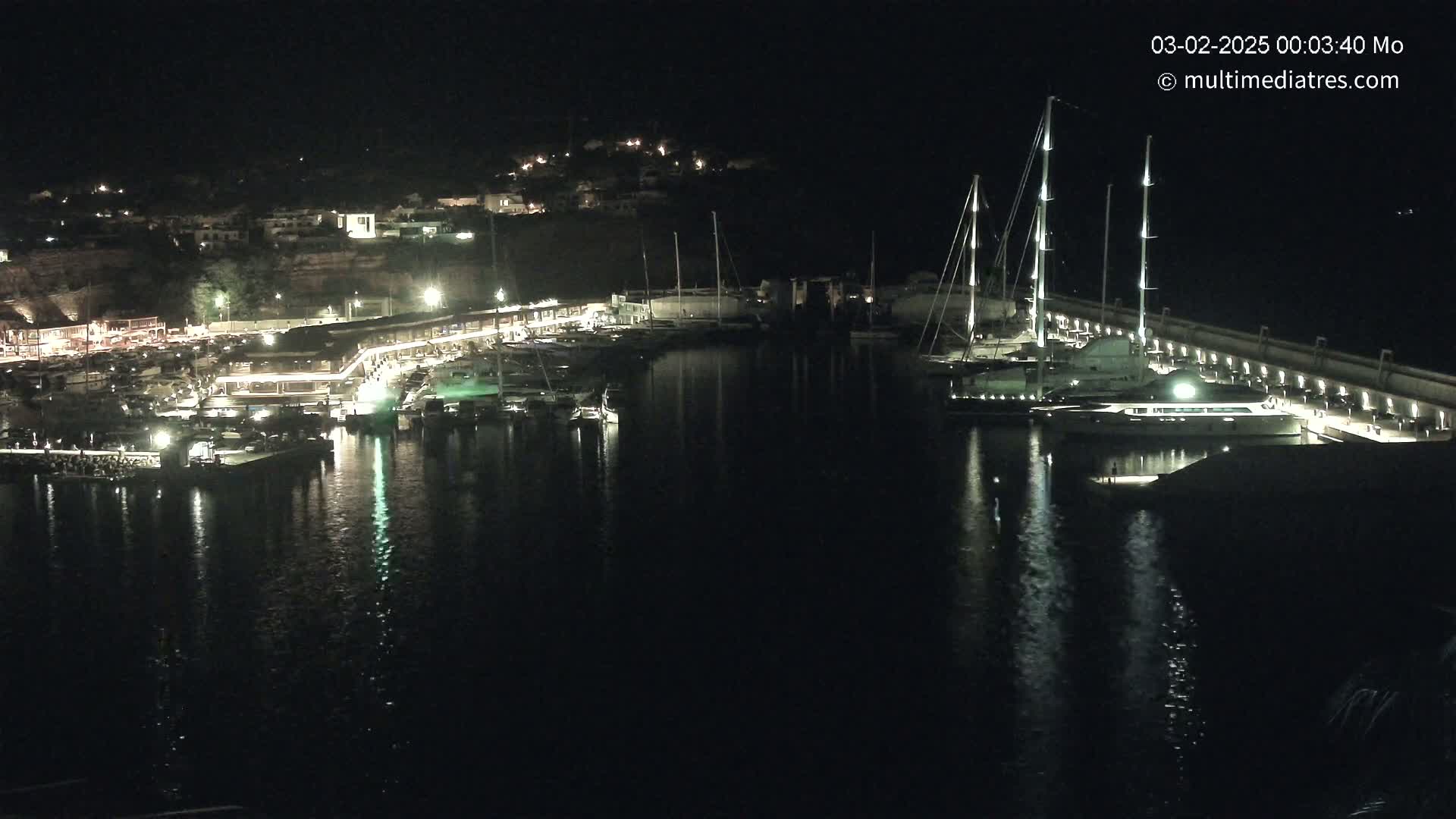 A marina at night, filled with boats and lit buildings, reflecting in calm water under a dark sky.