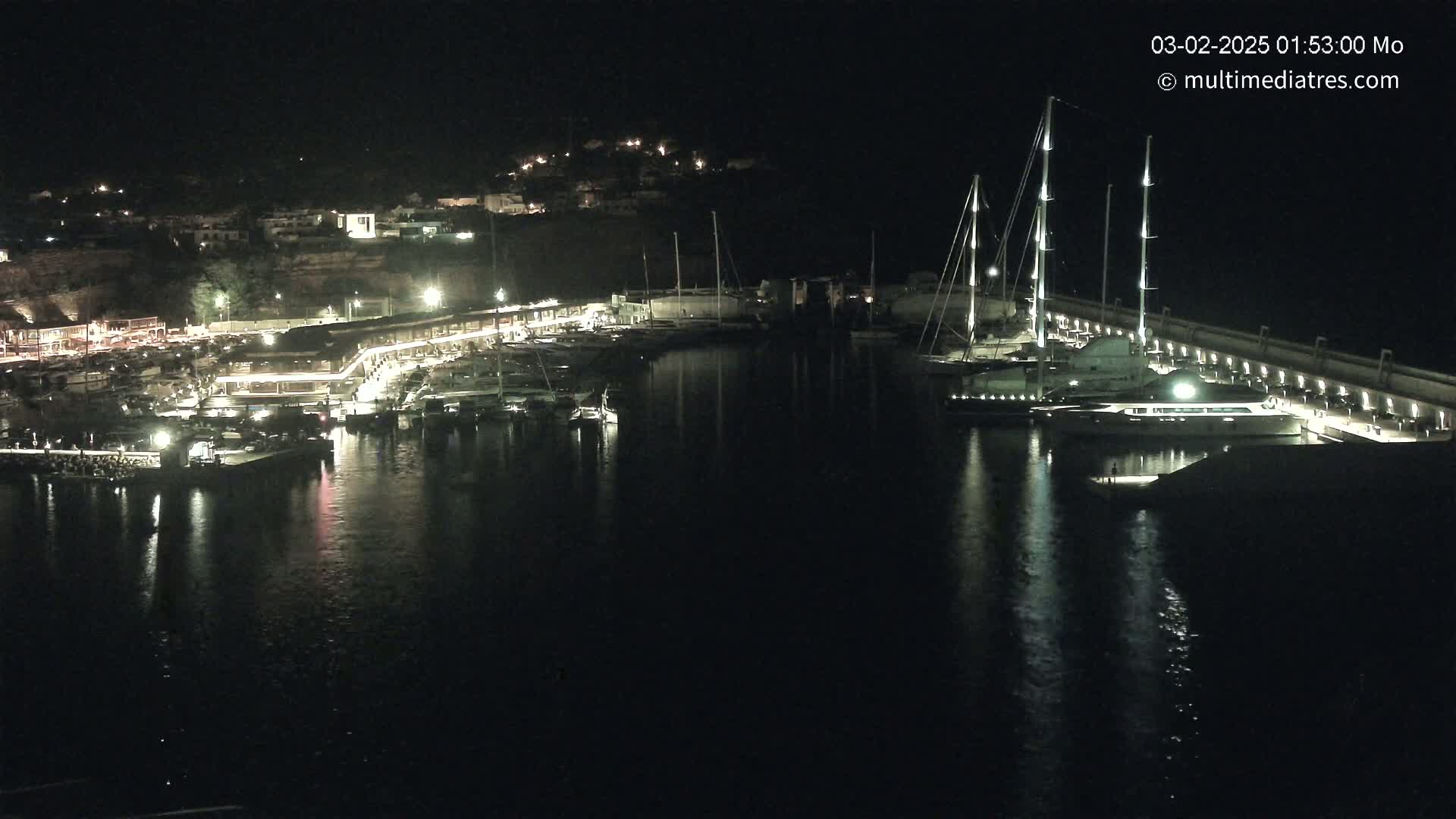 A nighttime harbor scene shows numerous boats docked along a lit waterfront, with a larger yacht prominently featured near a seawall under a clear, dark sky.