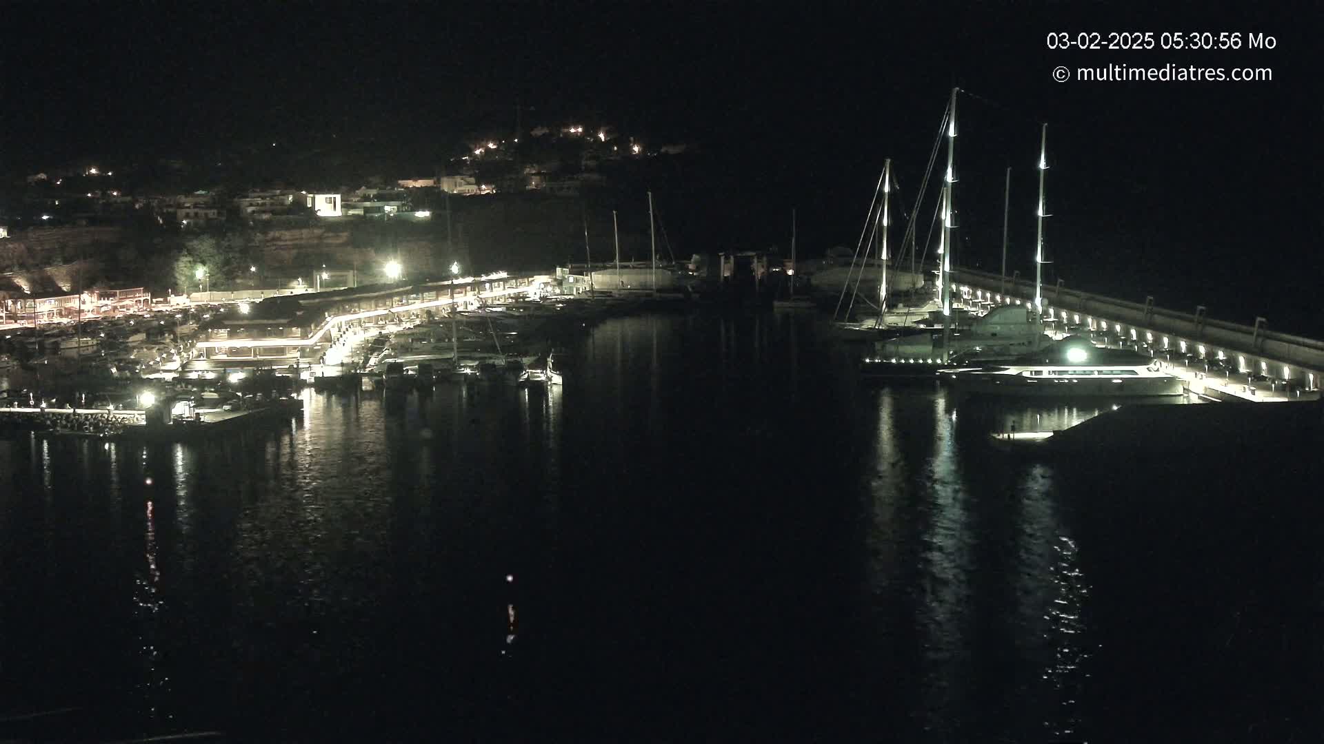 A nighttime view of a harbor filled with boats, docked along a lit waterfront with a hill in the background.