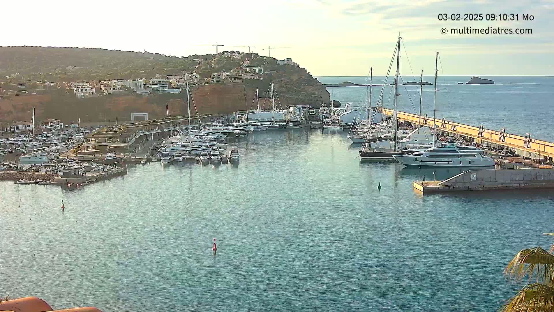 A calm harbor filled with numerous yachts and sailboats is nestled against a rocky hillside under a clear sky.
