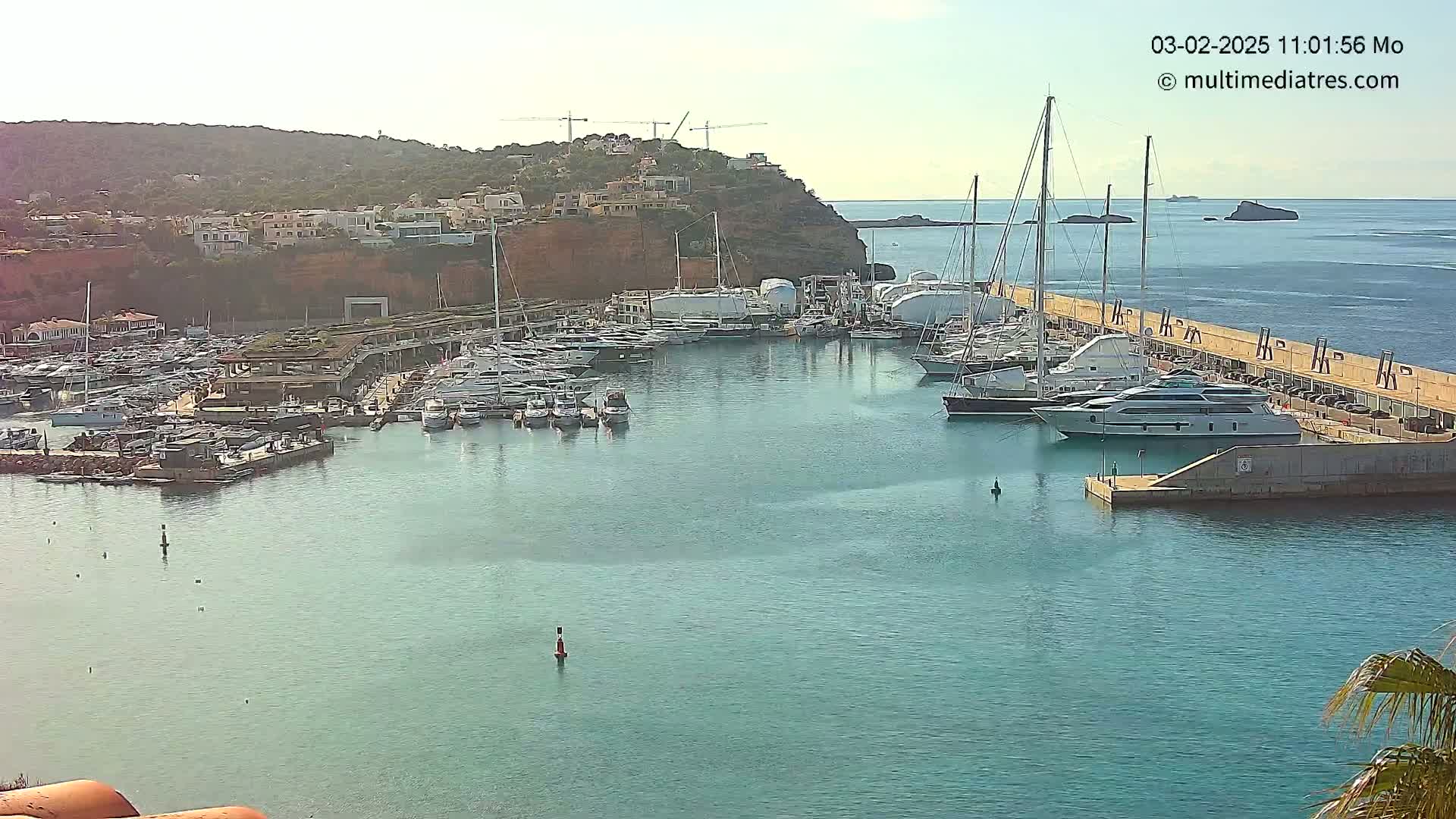 A marina filled with numerous yachts and sailboats is situated next to a hilltop town under a clear sky.