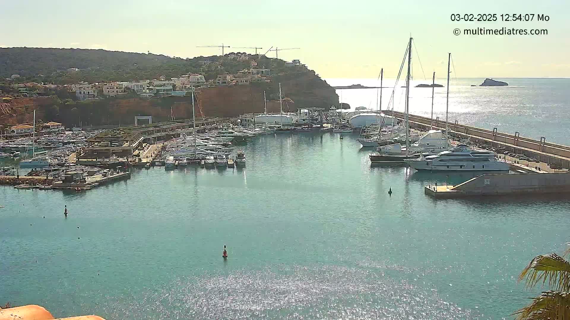 A marina filled with numerous yachts and boats is situated on calm, sunny water next to a hillside with buildings and construction cranes.