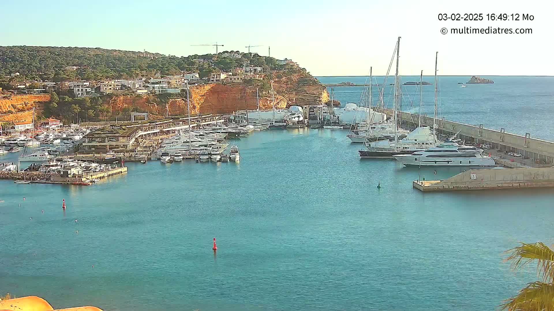 A marina filled with numerous yachts and boats is situated on calm, clear water, against a backdrop of a hillside town under a sunny sky.