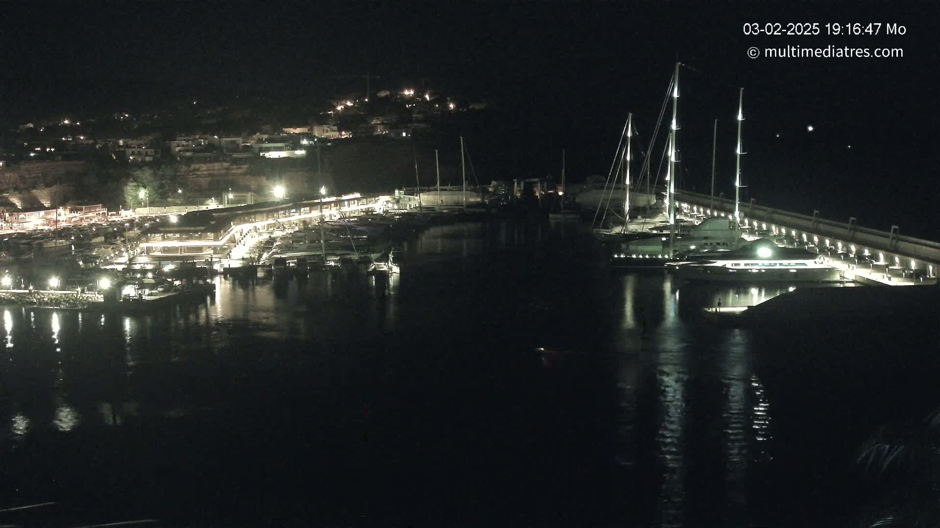 A nighttime view of a marina filled with boats, illuminated by lights, with calm water reflecting the lights.