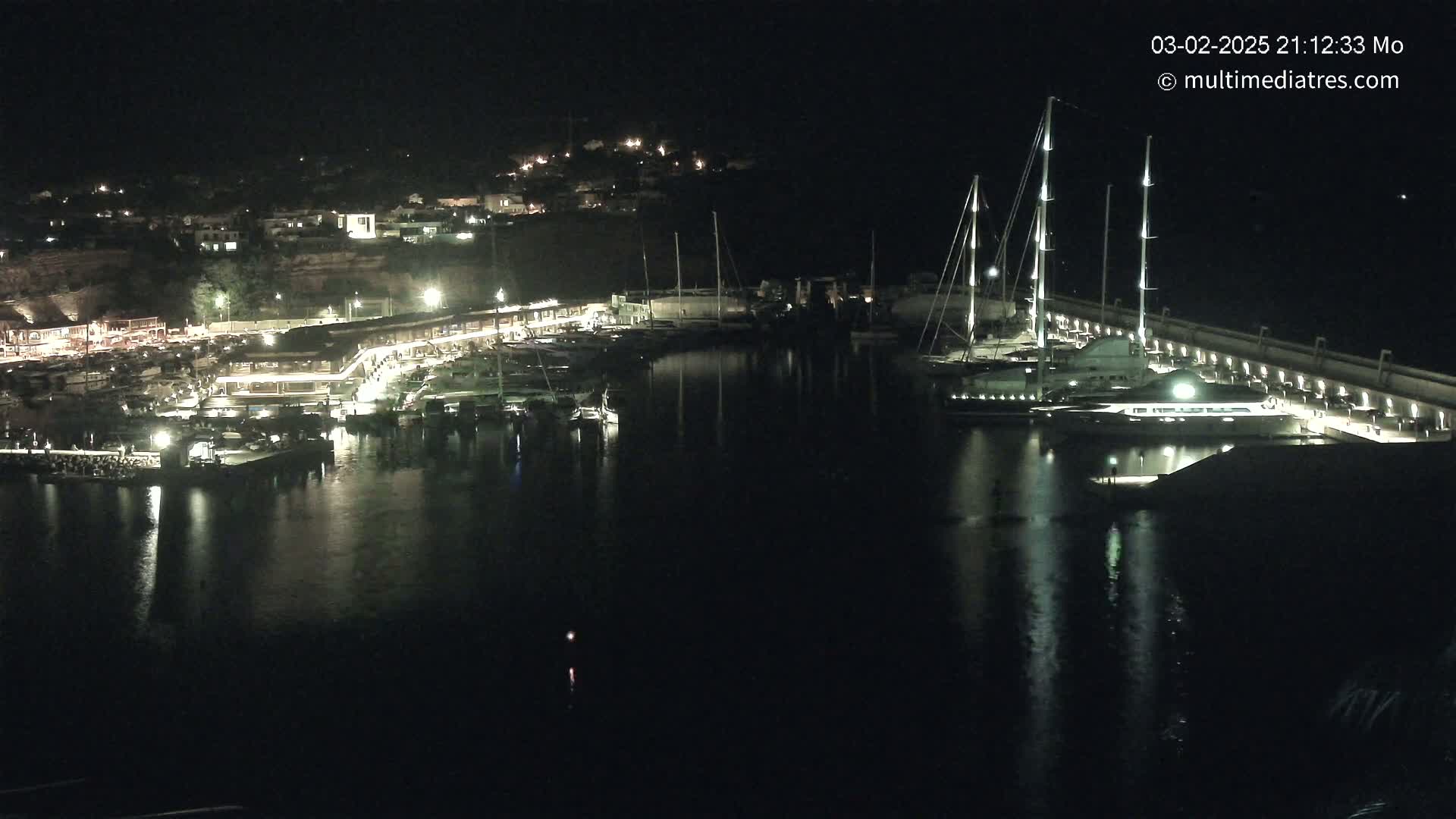 A nighttime view of a harbor filled with boats and yachts, with a town illuminated on a hillside in the background.