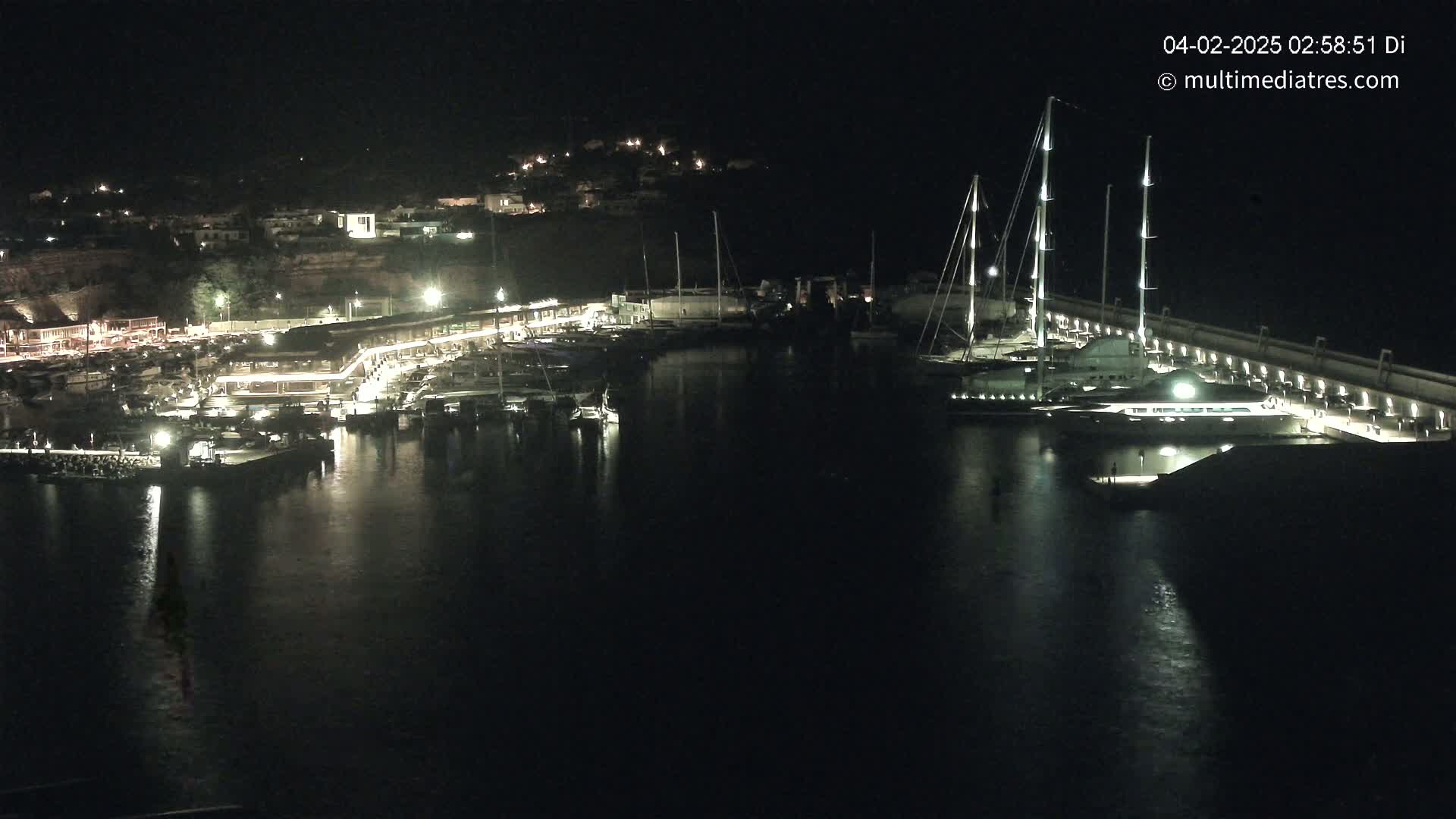 A marina at night, filled with boats and illuminated by lights, sits next to a town nestled on a hillside.