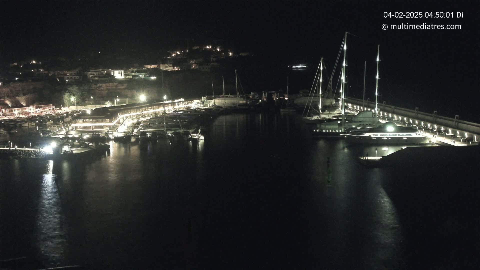 A marina at night, filled with docked boats and yachts, is illuminated by lights along a waterfront walkway under a dark sky.