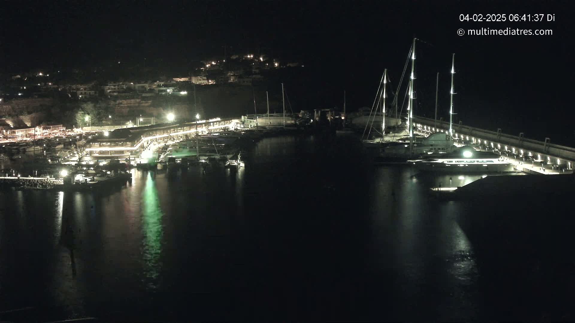 A nighttime view of a harbor filled with boats, illuminated by lights along a waterfront, under a clear, dark sky.