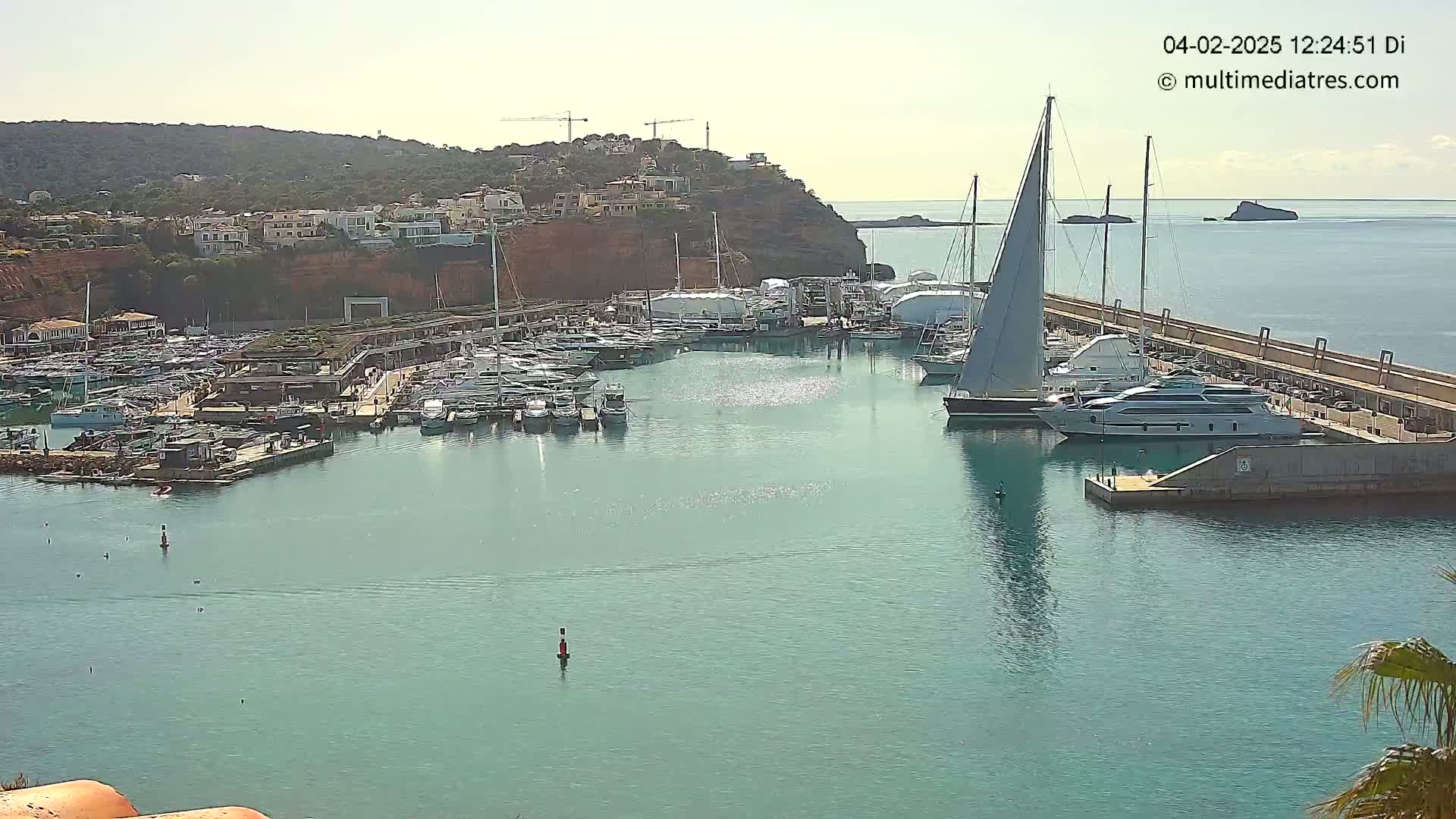 A marina filled with numerous yachts and sailboats is situated on calm, teal water next to a hillside of buildings under a bright, sunny sky.