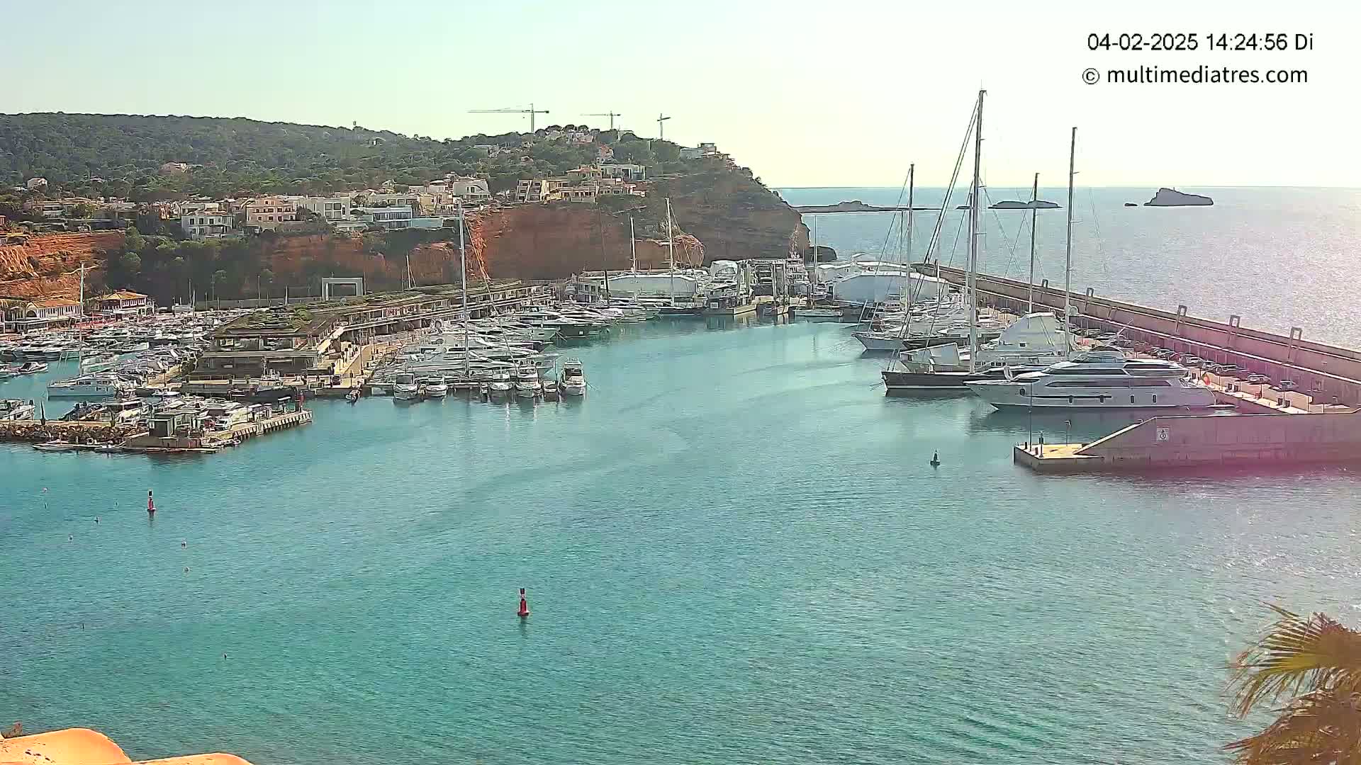 A marina filled with numerous yachts and boats is nestled into a calm, clear body of water beside a red-toned cliff with buildings atop it under a sunny sky.
