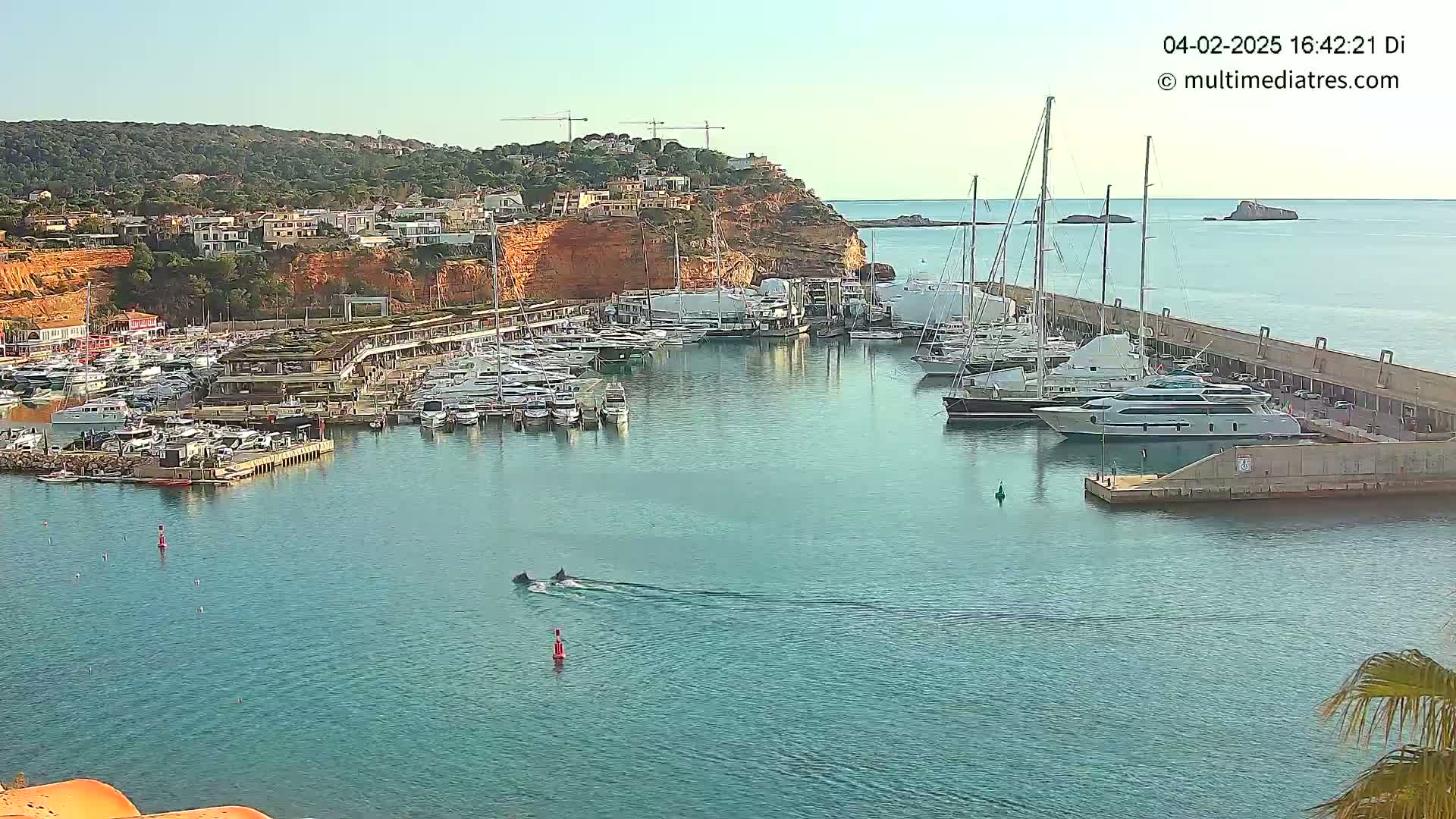 A marina filled with numerous boats and yachts is situated against a backdrop of a coastal town nestled on a reddish cliff, under a clear, sunny sky.