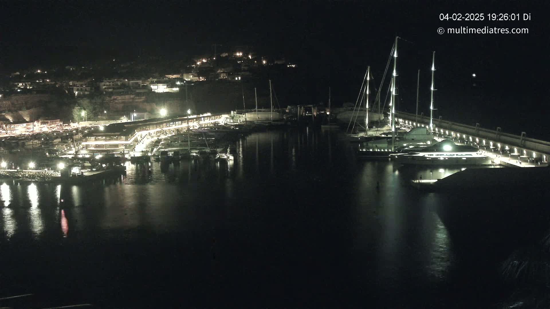 A nighttime view of a marina filled with boats, illuminated by lights, against a dark backdrop of a hillside town.