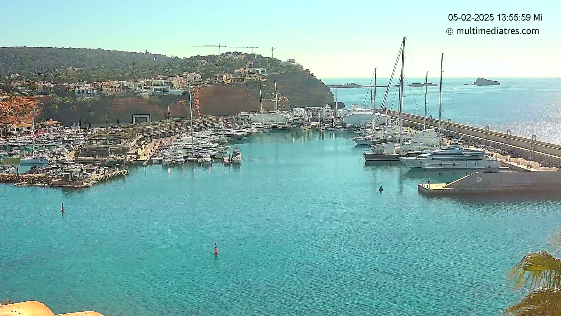 A marina filled with numerous yachts and boats is situated on a calm, clear blue body of water next to a red-clay hillside on a sunny day.