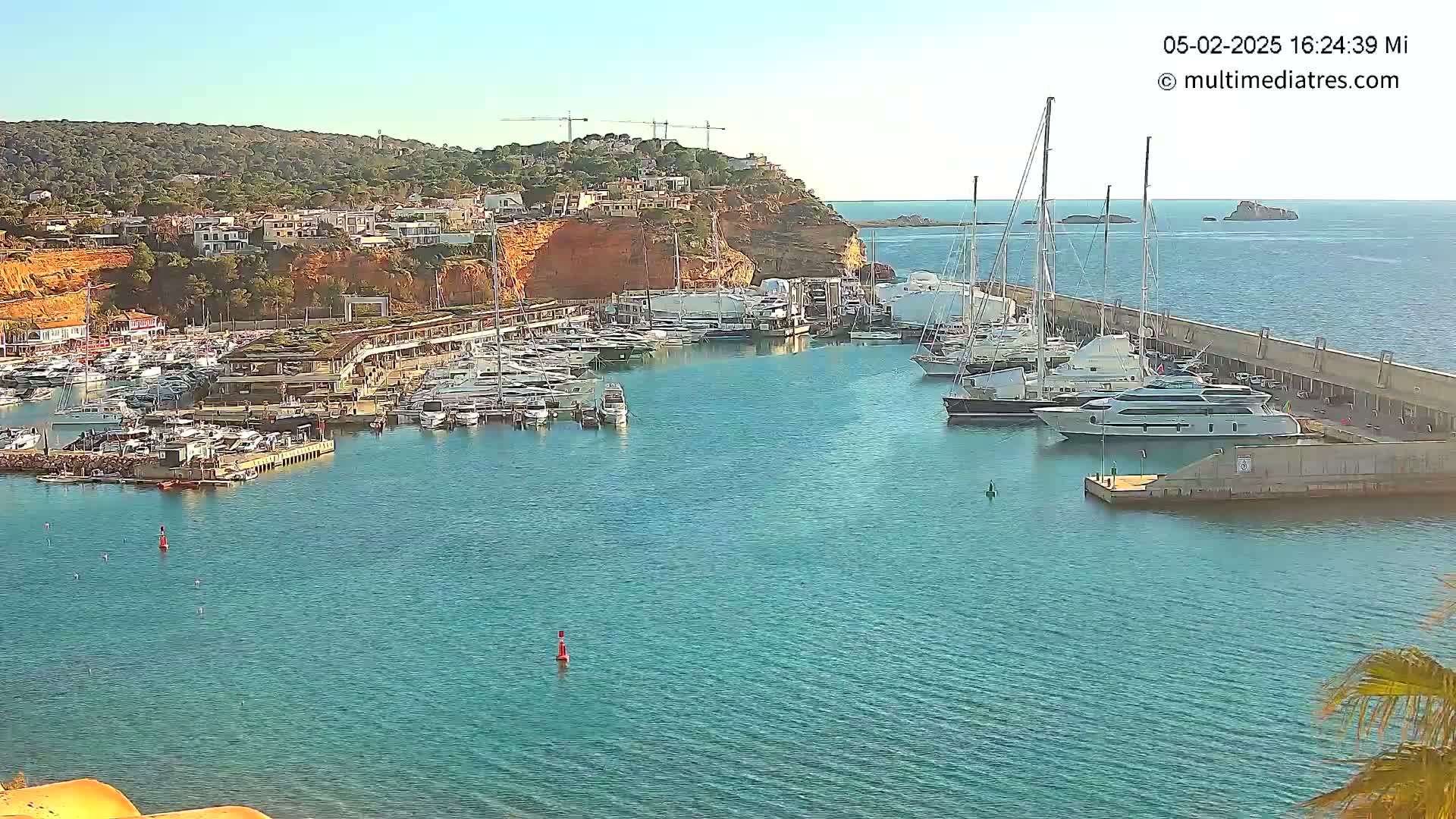 A marina filled with yachts and sailboats is nestled in a calm, blue bay, under a clear sunny sky, with a hillside town visible in the background.