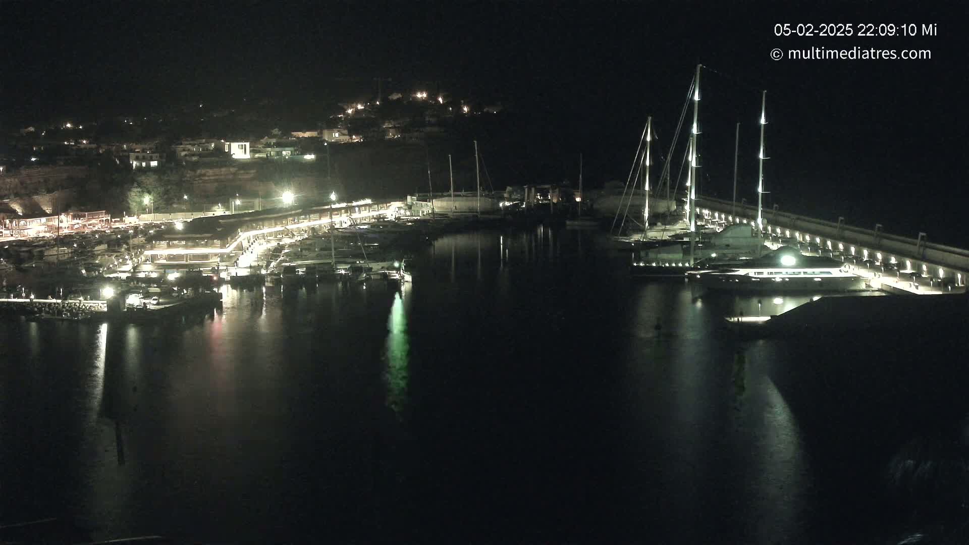 A nighttime view of a harbor filled with boats and yachts, illuminated by lights from the waterfront buildings and docked vessels.