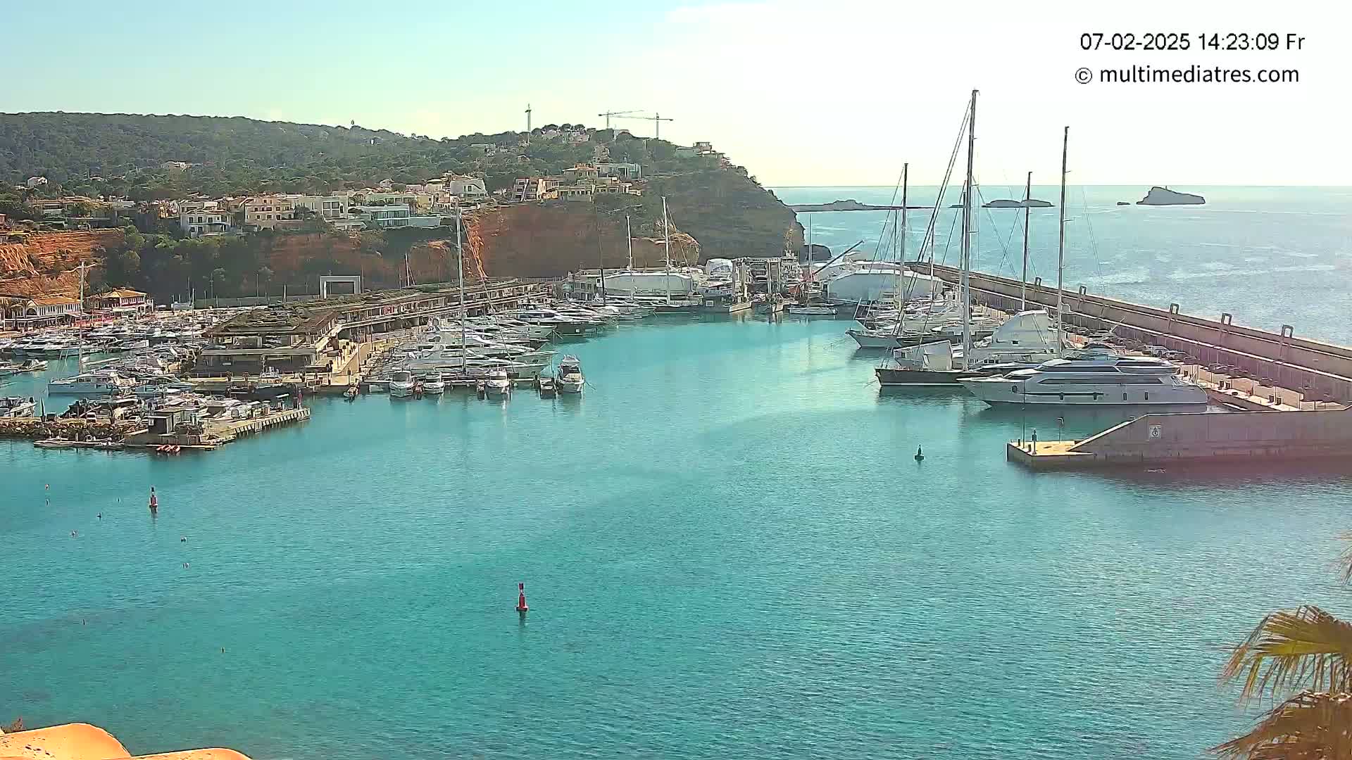 A marina filled with numerous yachts and boats is situated on a calm, turquoise body of water next to a hillside town under a clear, sunny sky.
