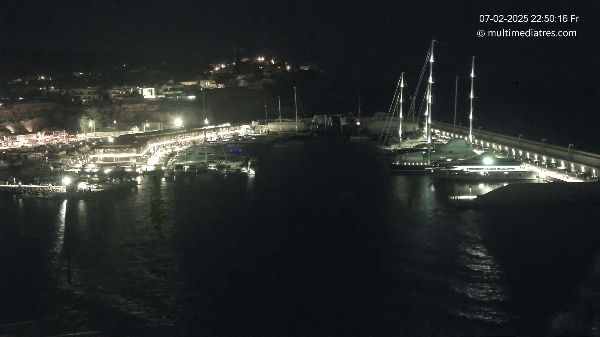 A nighttime view of a marina filled with boats, docked along a lit waterfront and hillside buildings.