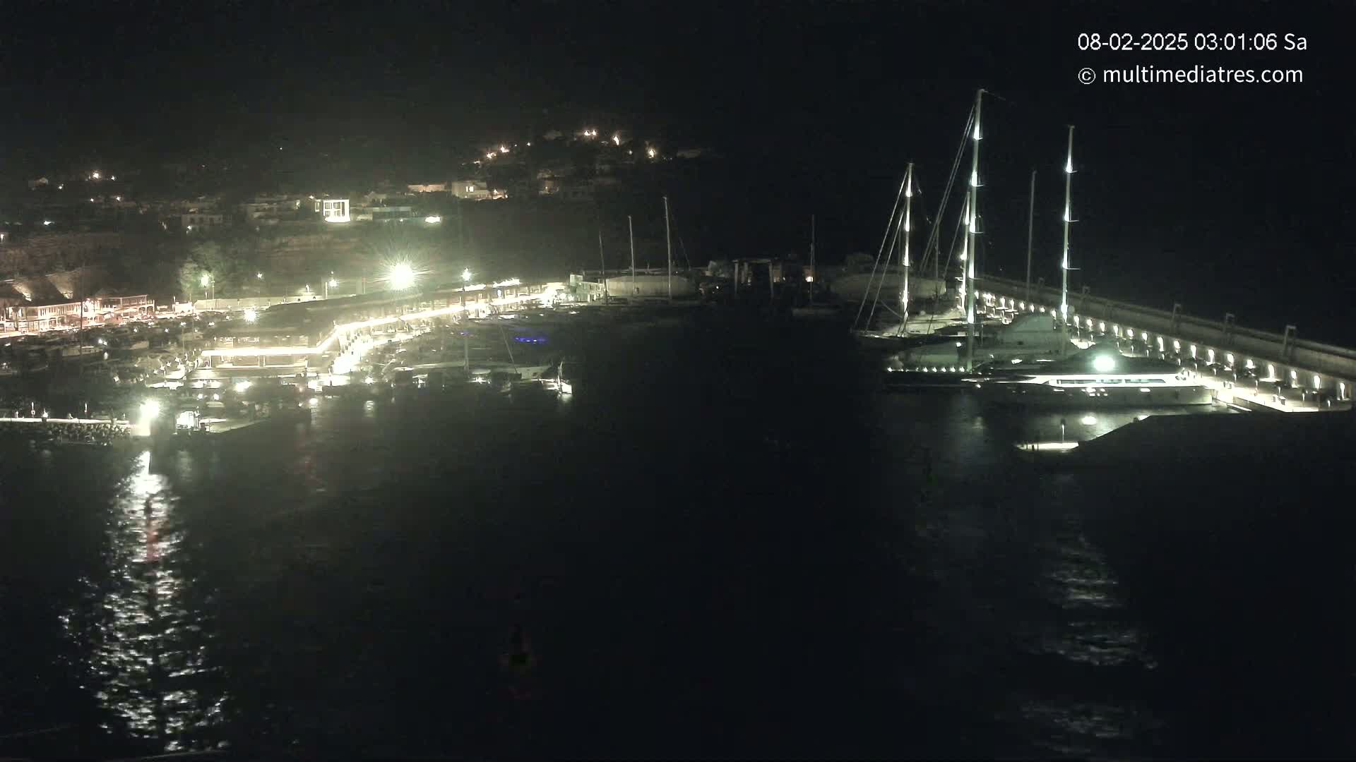 A nighttime aerial view of a marina with several boats docked, illuminated buildings along the shoreline, and calm water.