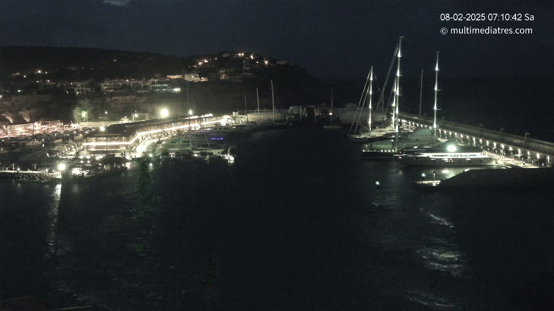 A nighttime view of a harbor filled with boats and yachts, illuminated by lights along a waterfront building and a seawall.