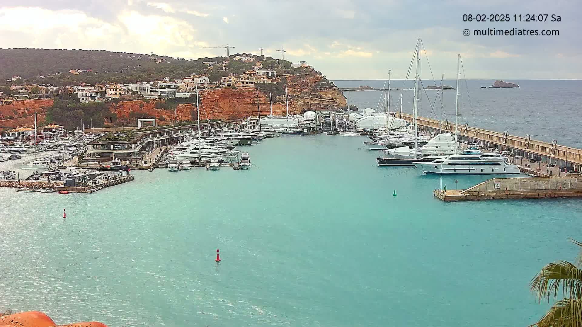 A marina filled with yachts and sailboats is situated in a calm, turquoise bay under a partly cloudy sky, with a hillside of buildings visible in the background.