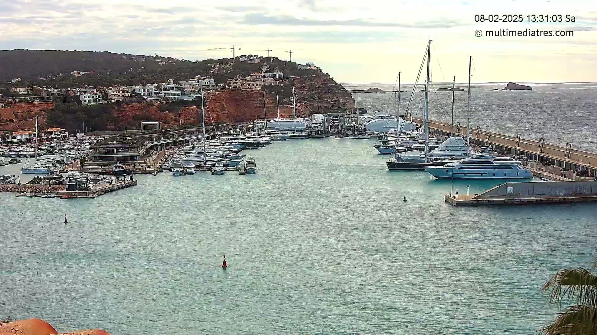 A marina filled with many yachts and boats is nestled against a rocky hillside under a cloudy sky.