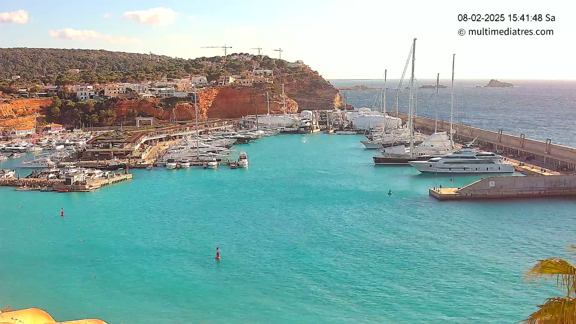 A marina filled with yachts and boats is nestled into a turquoise bay, backed by a hillside of buildings under a mostly clear sky.