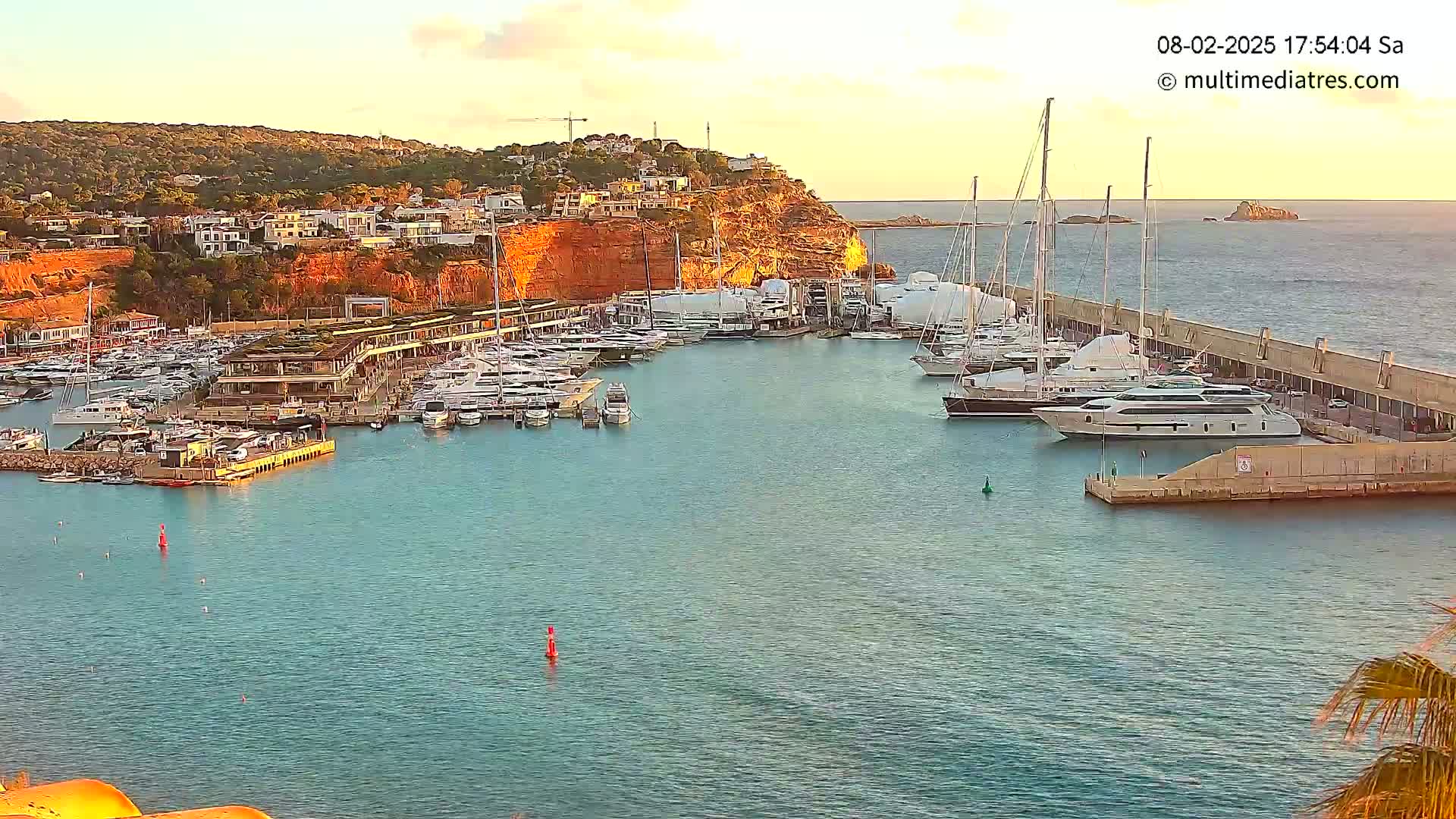 A calm harbor filled with numerous yachts and boats is nestled against a rocky hillside dotted with buildings, under a clear, sunset sky.
