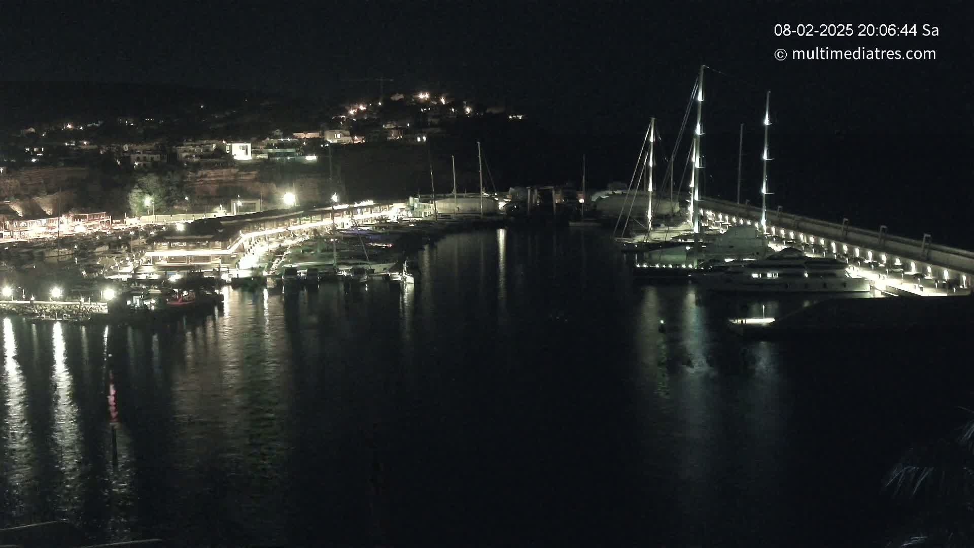 A nighttime view of a marina filled with yachts and boats, illuminated by lights along the docks and buildings on the shore.