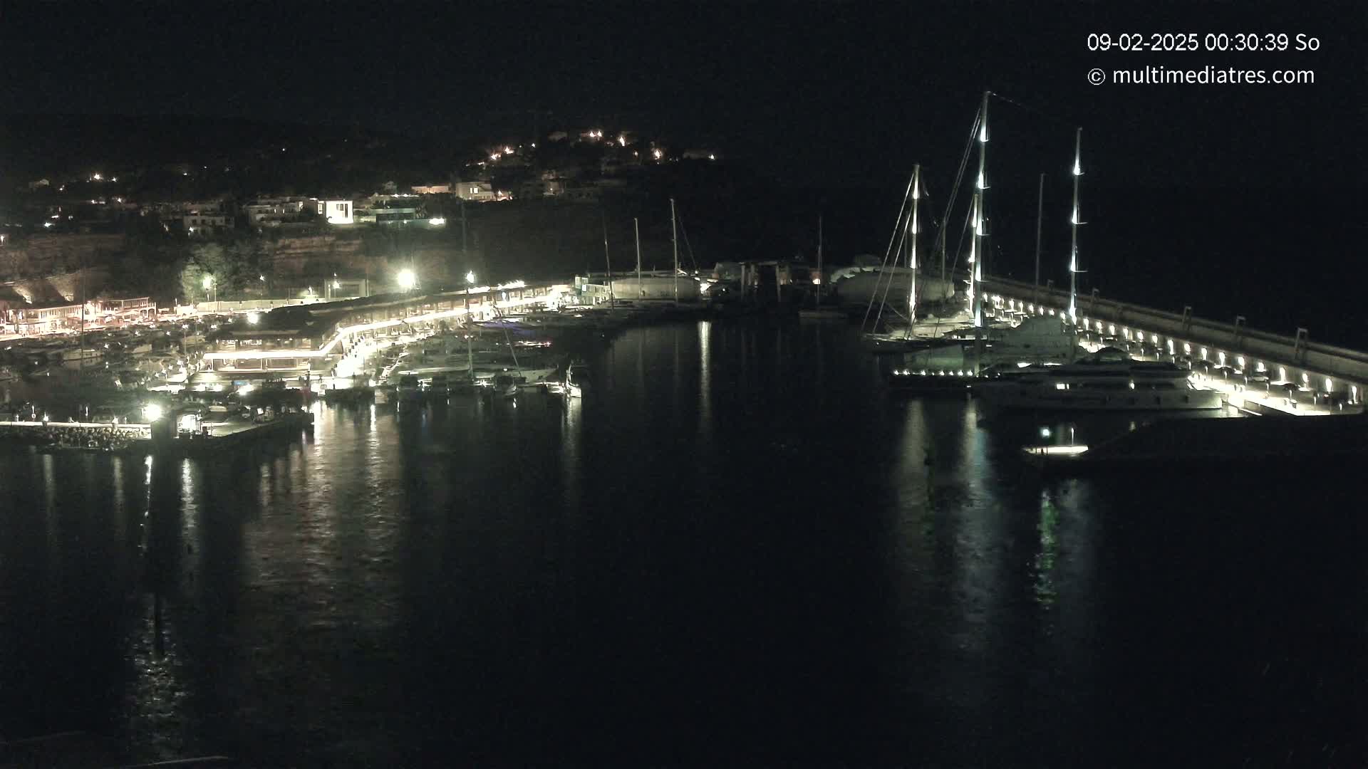A nighttime view of a harbor filled with boats, illuminated by lights along a waterfront with buildings visible in the background.