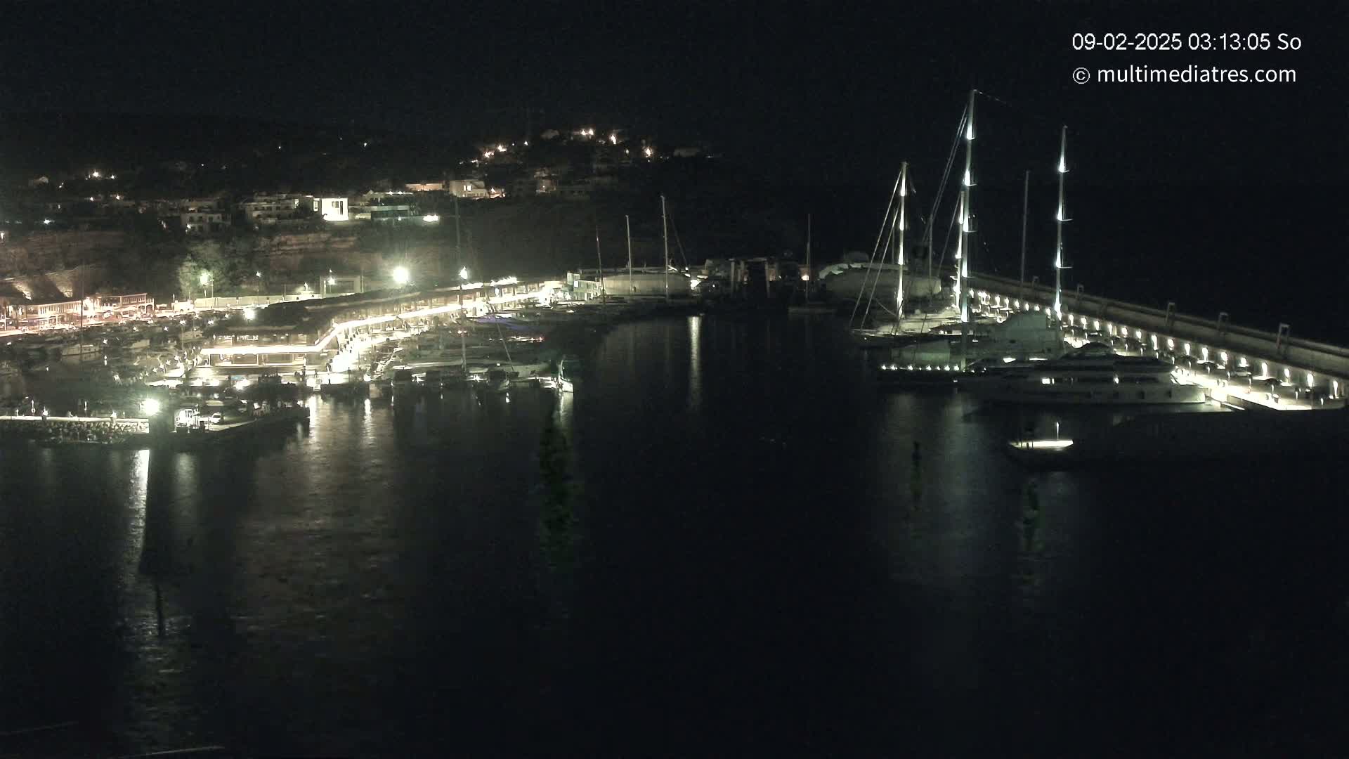A nighttime view of a marina filled with boats, illuminated by lights along the docks and buildings on the shore.