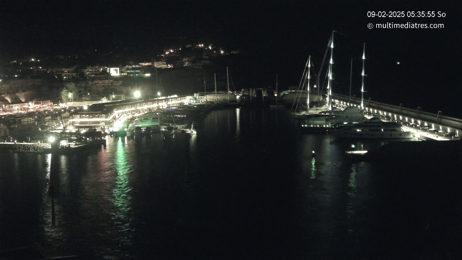 A nighttime view of a harbor filled with boats and yachts, illuminated by lights along a waterfront.