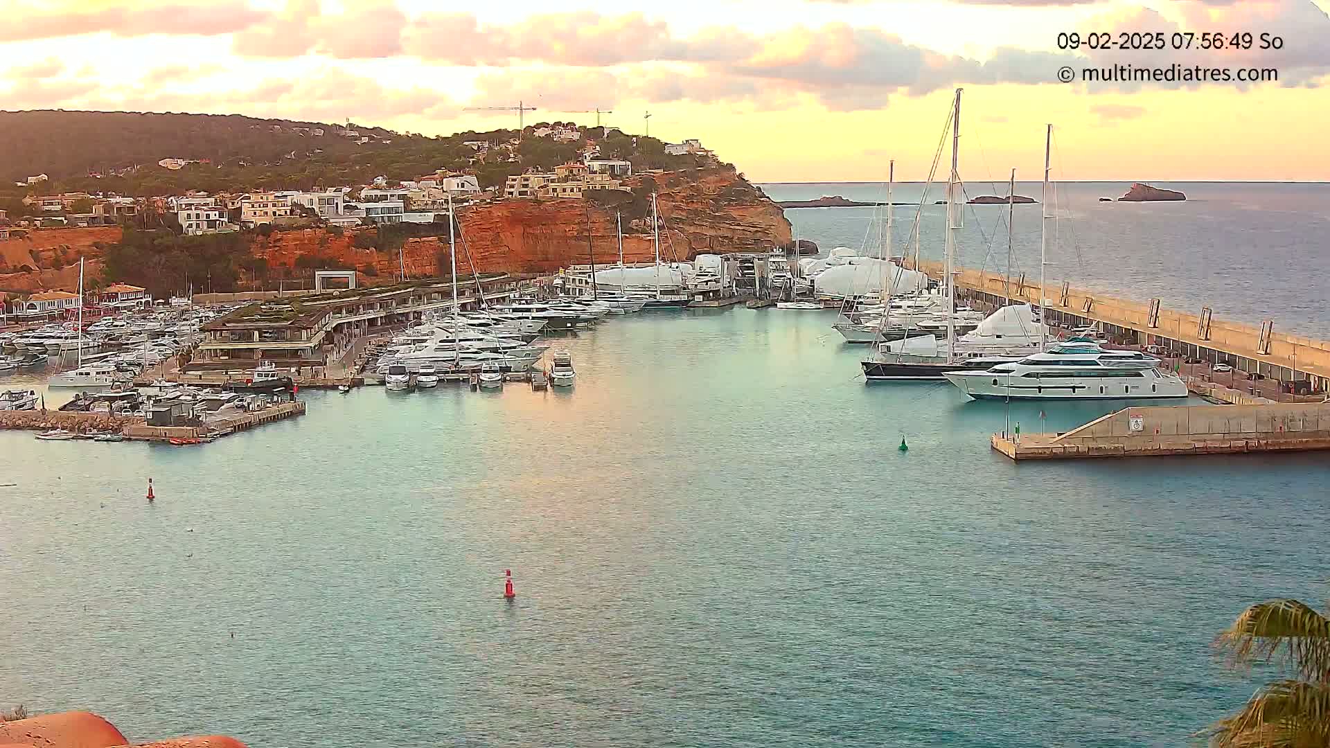 A marina filled with numerous yachts and boats is situated along a rocky coast under a partly cloudy, sunset sky.
