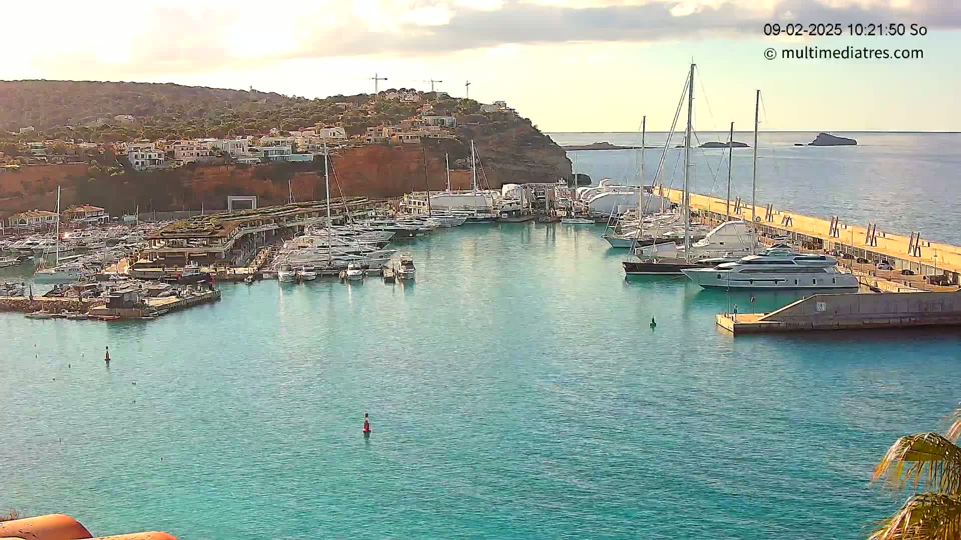 A marina filled with numerous yachts and boats is nestled against a red cliffside under a partly cloudy sky.