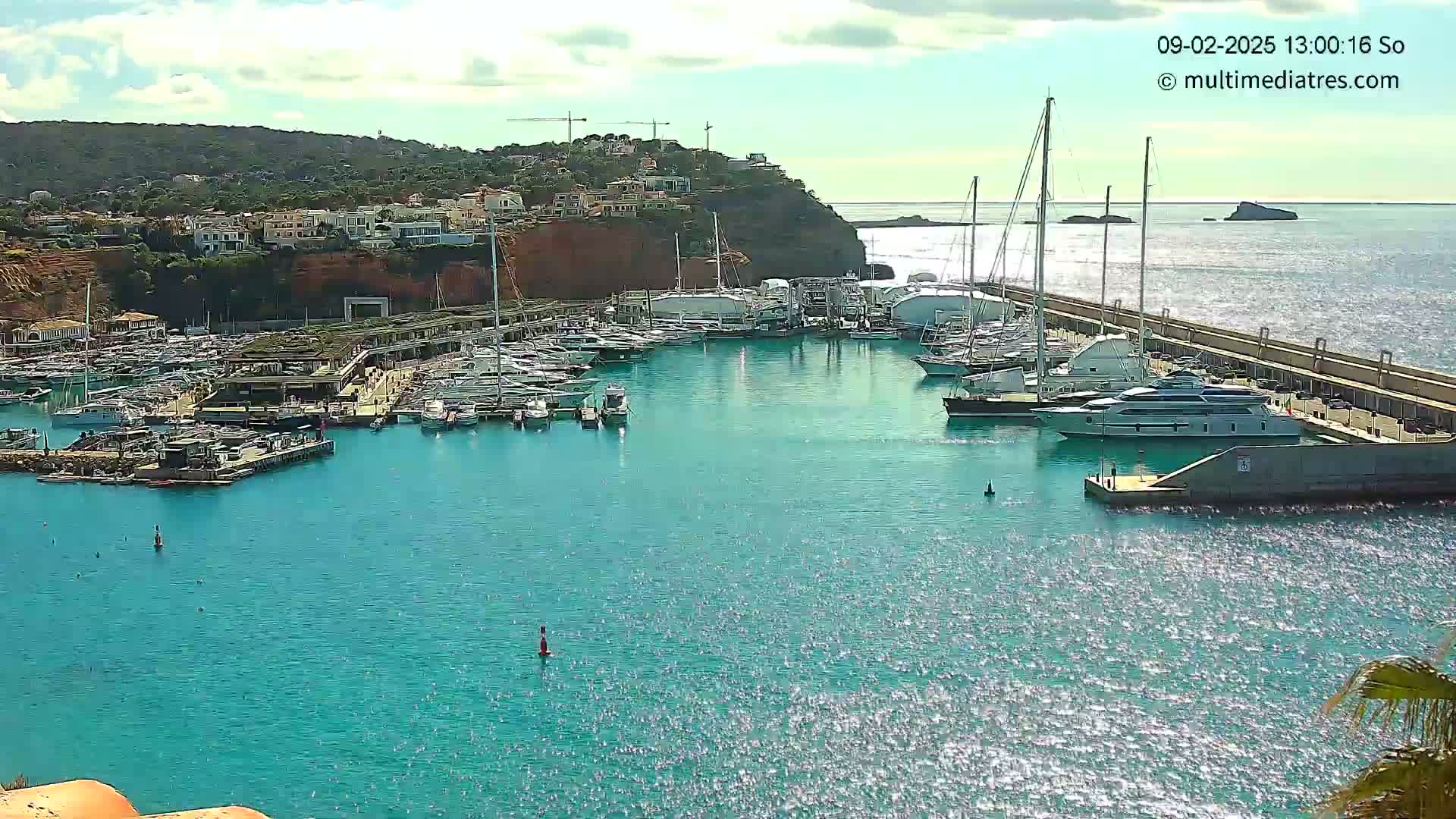A marina filled with yachts and boats is nestled into a coastline under a partly cloudy sky.